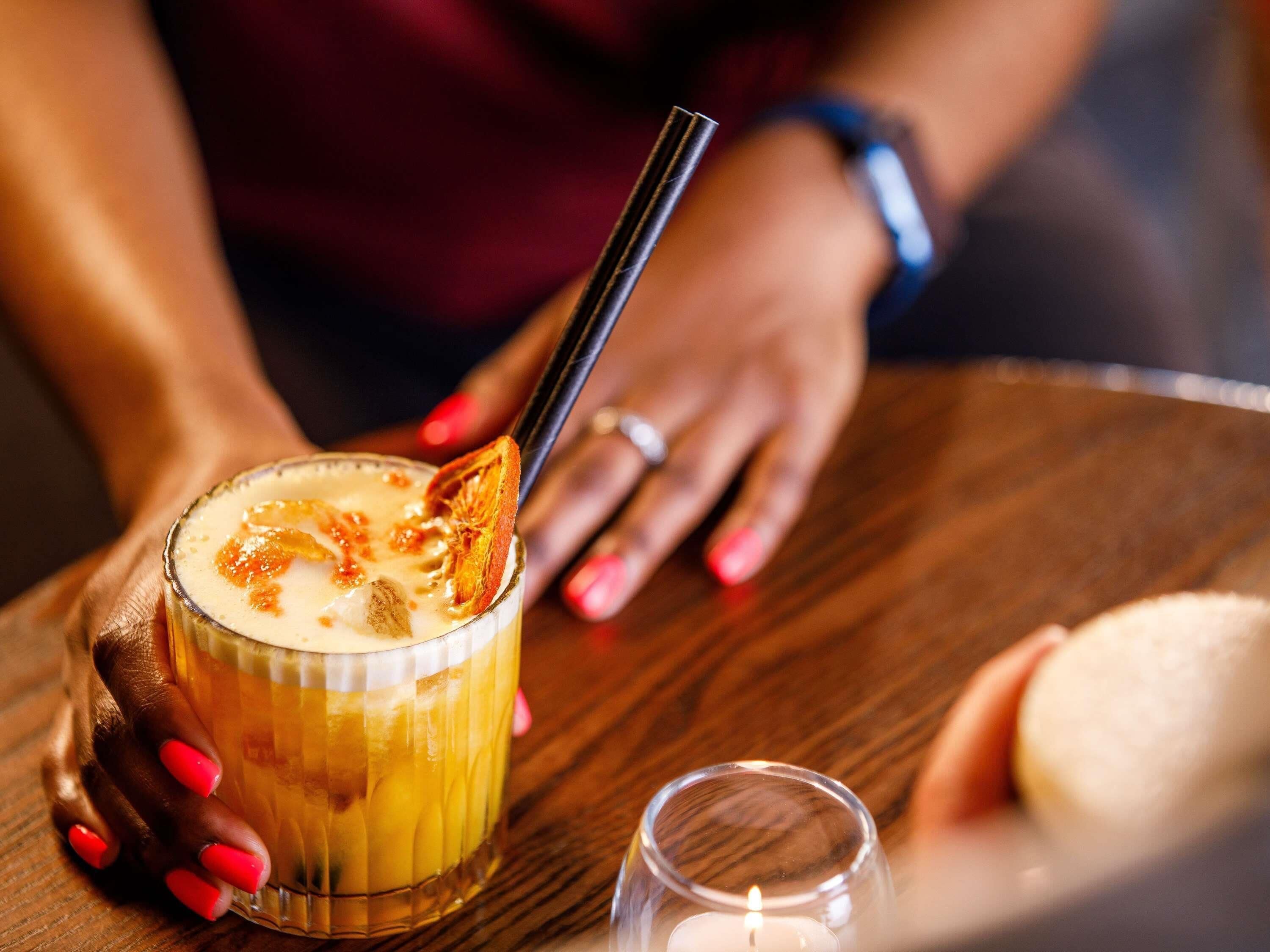 A person holding a glass with a straw, a candle, and a dessert item on a wooden table.