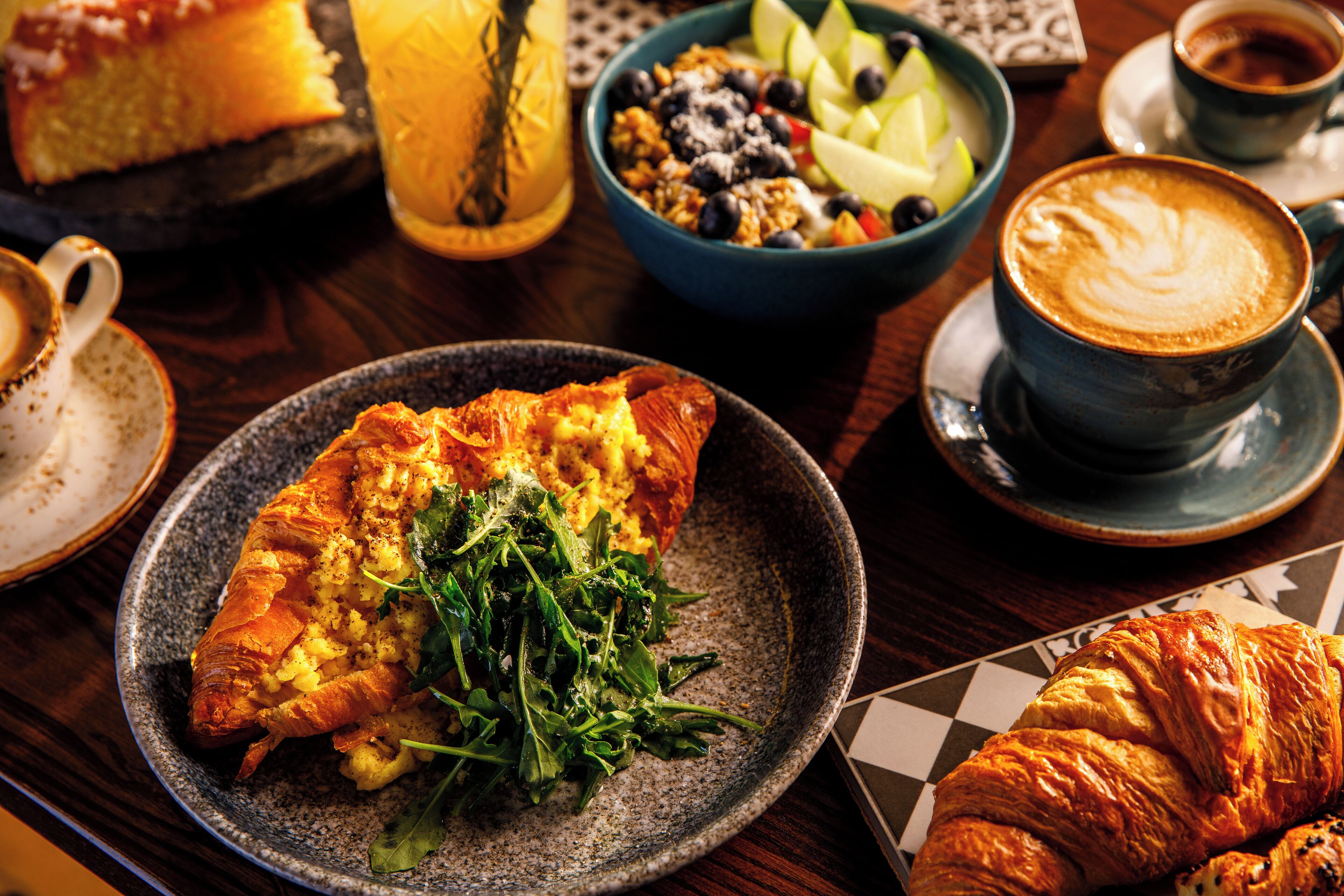 A dining table with a croissant, a bowl of fruit, a cup of coffee, and a slice of cake.