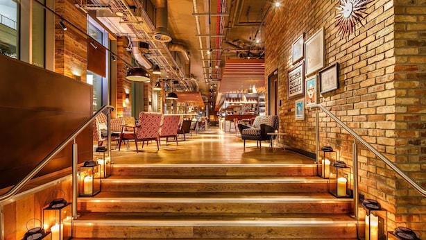 A staircase with wooden steps and lanterns, leading to a bar area with seating and a brick wall with a sunburst clock.