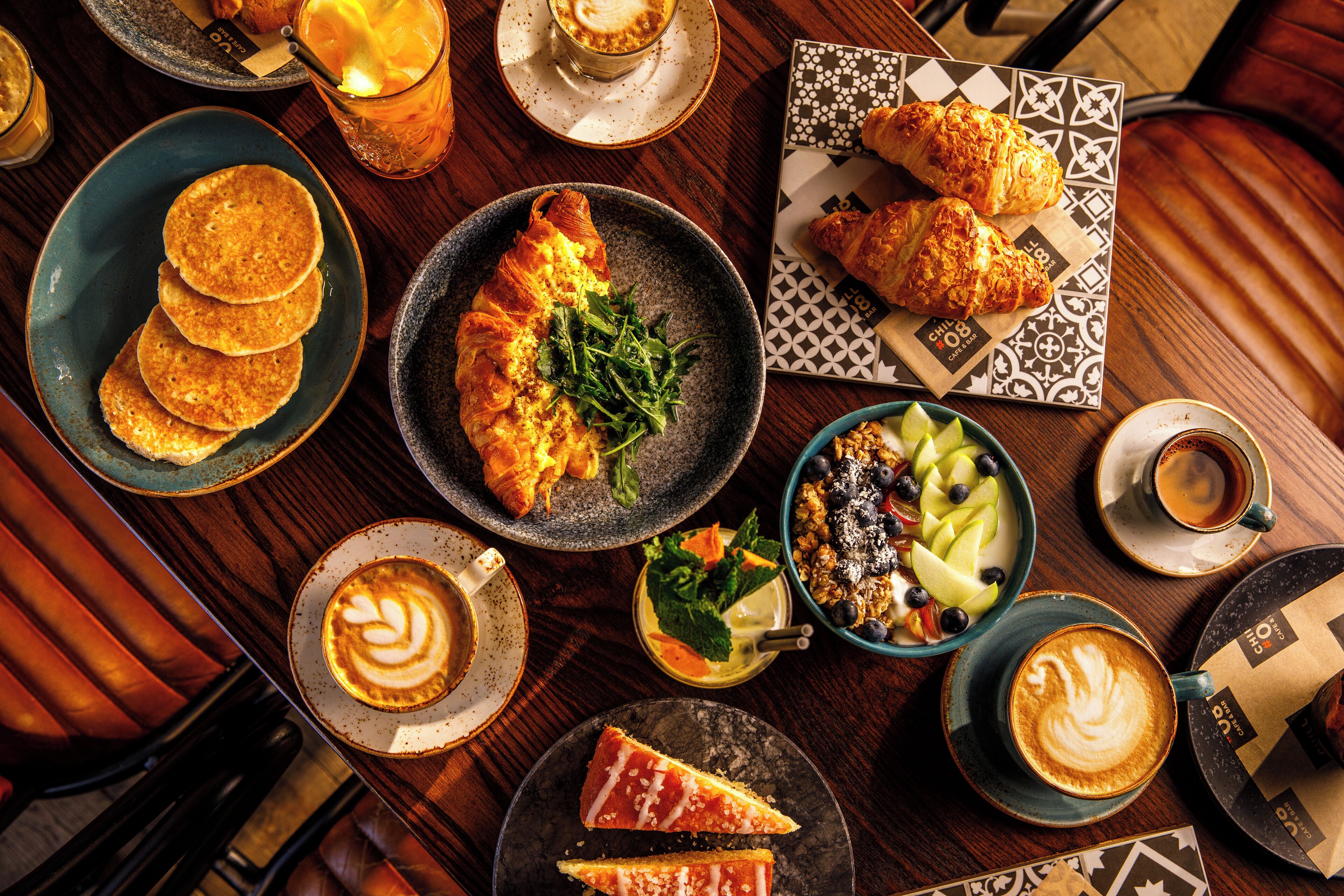 A table with various breakfast items including croissants, pancakes, and a croissant with greens. There are also cups of coffee and a bowl of fruit.