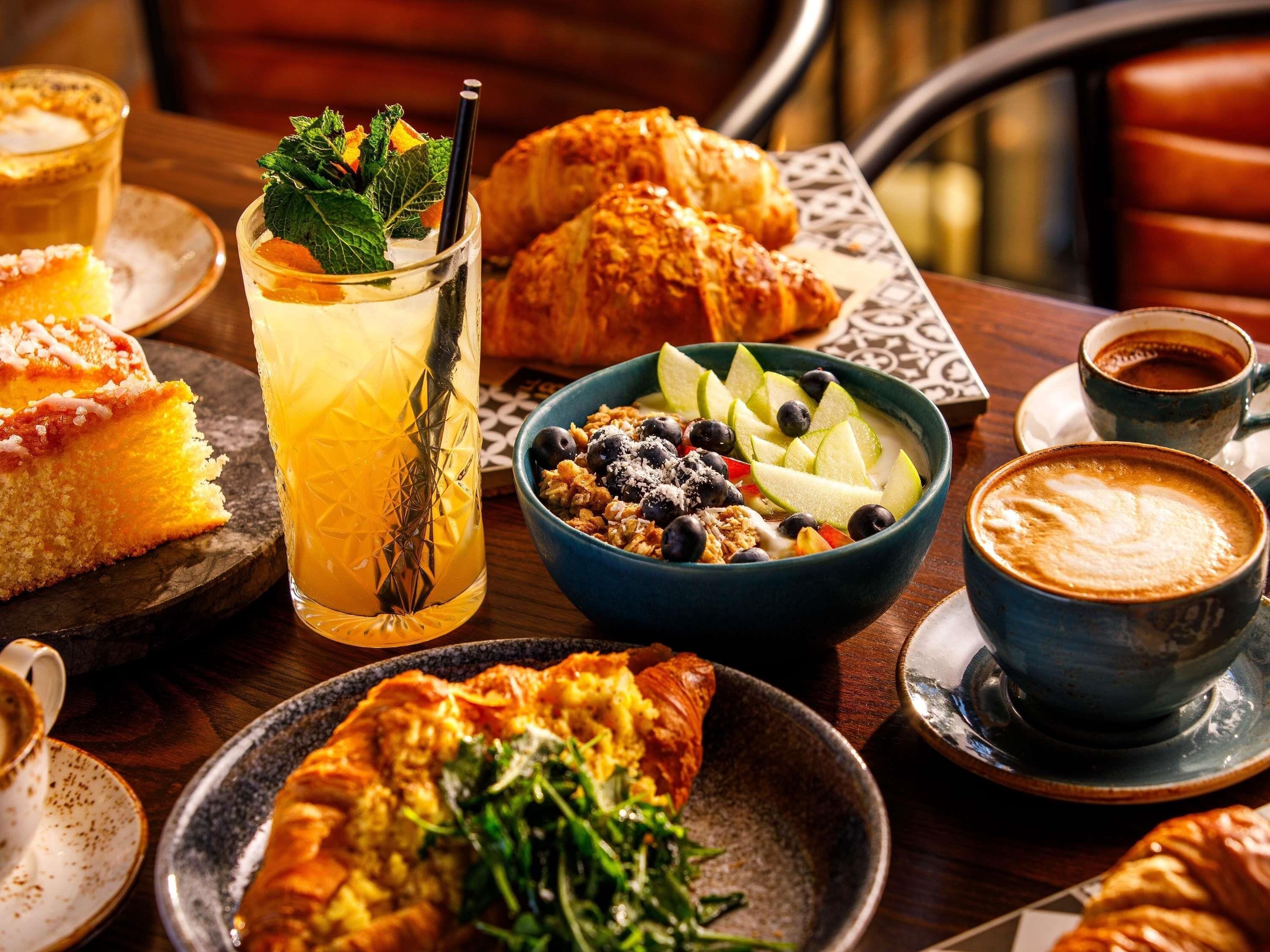 Una mesa con diversos alimentos, incluyendo pasteles, un bol de fruta, un vaso de bebida helada y una taza de café.