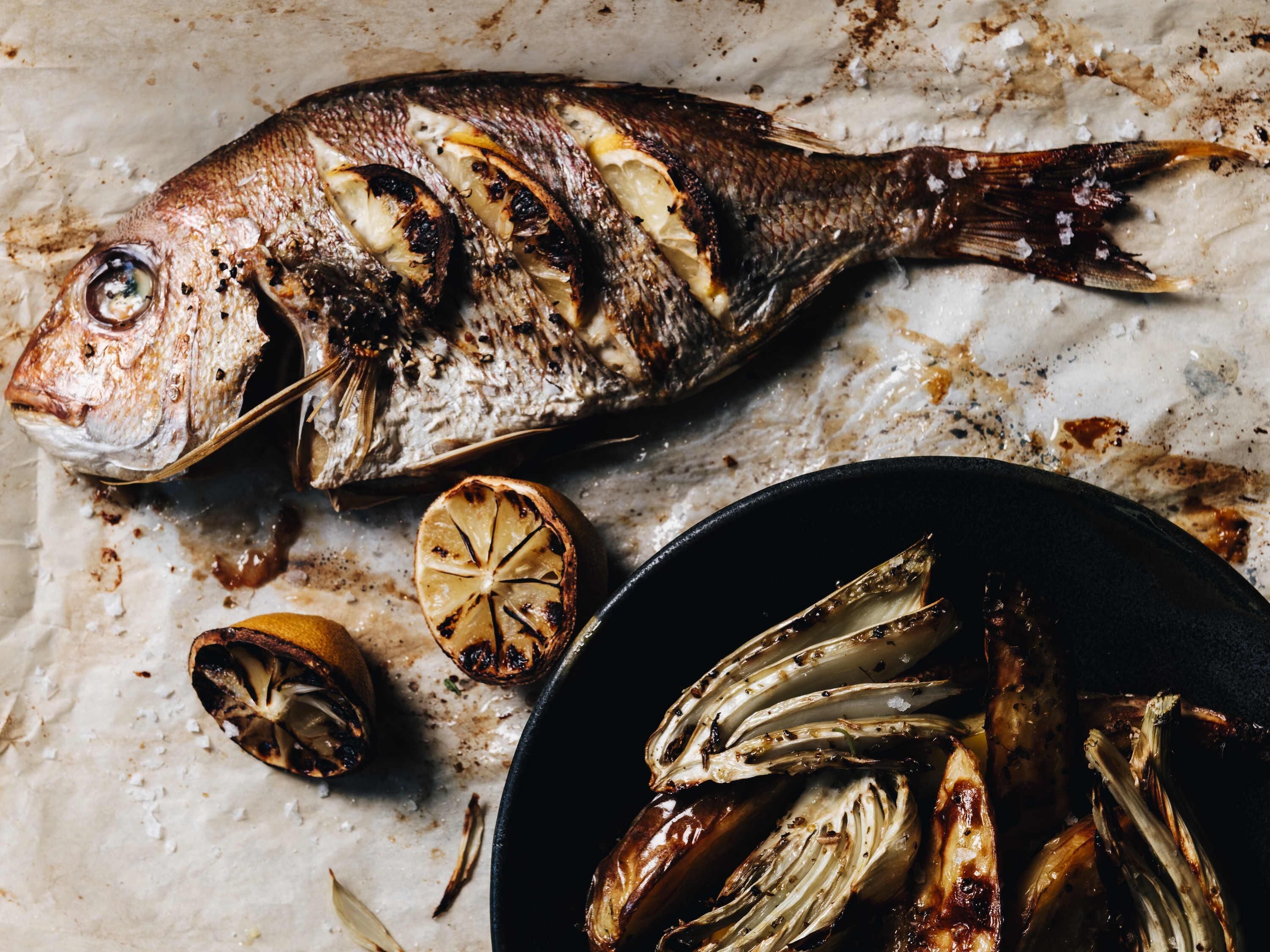 A roasted fish with lemon slices and grilled vegetables on a marble surface.