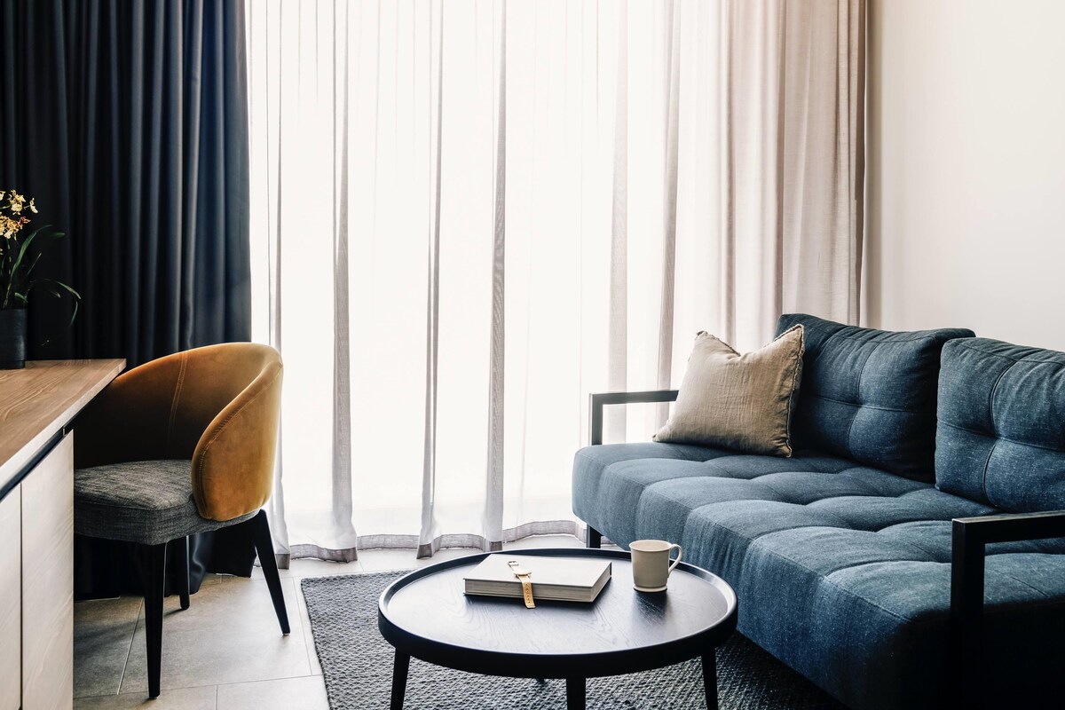 Un salon moderne avec un canapé, un fauteuil, une table basse et une vue dégagée à travers des rideaux légers.