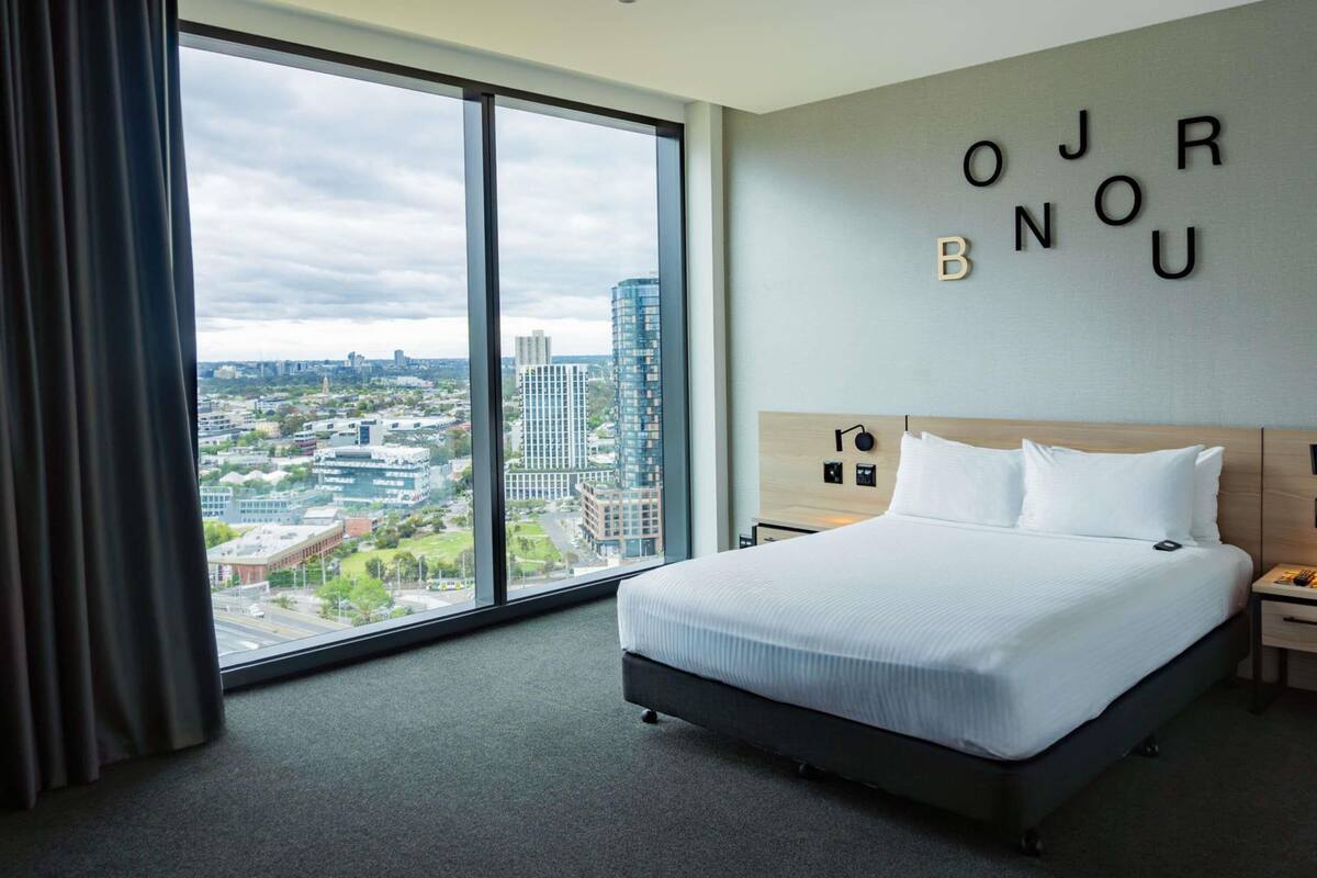 Quarto de hotel com uma grande janela, uma cama, uma mesa de cabeceira e uma parede com as letras que formam 'OJRBNOU'.
