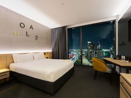Habitación de hotel con cama, escritorio, silla y una ventana grande con vista a la ciudad.