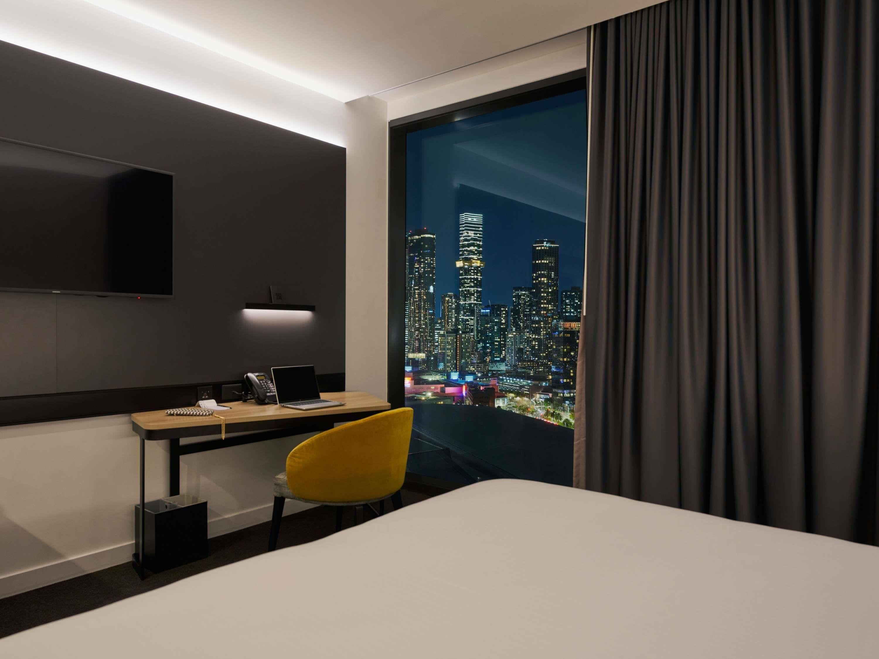 A hotel room with a desk, chair, and a large window with a cityscape view.