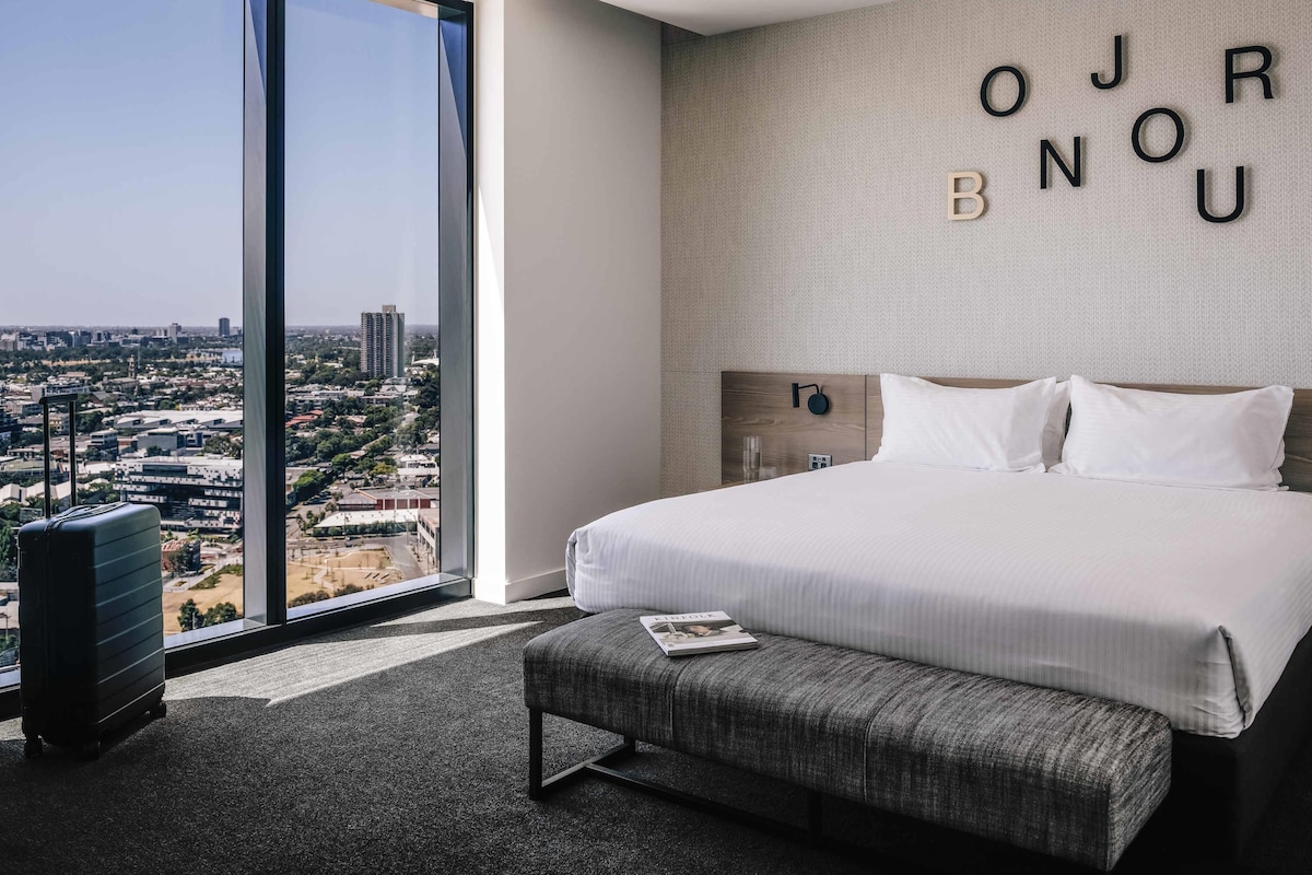 A hotel room with a large bed, a bench, a suitcase, and a view of a cityscape through a large window.