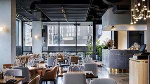A modern restaurant with tables set for dining, a bar area, and large windows offering a city view.