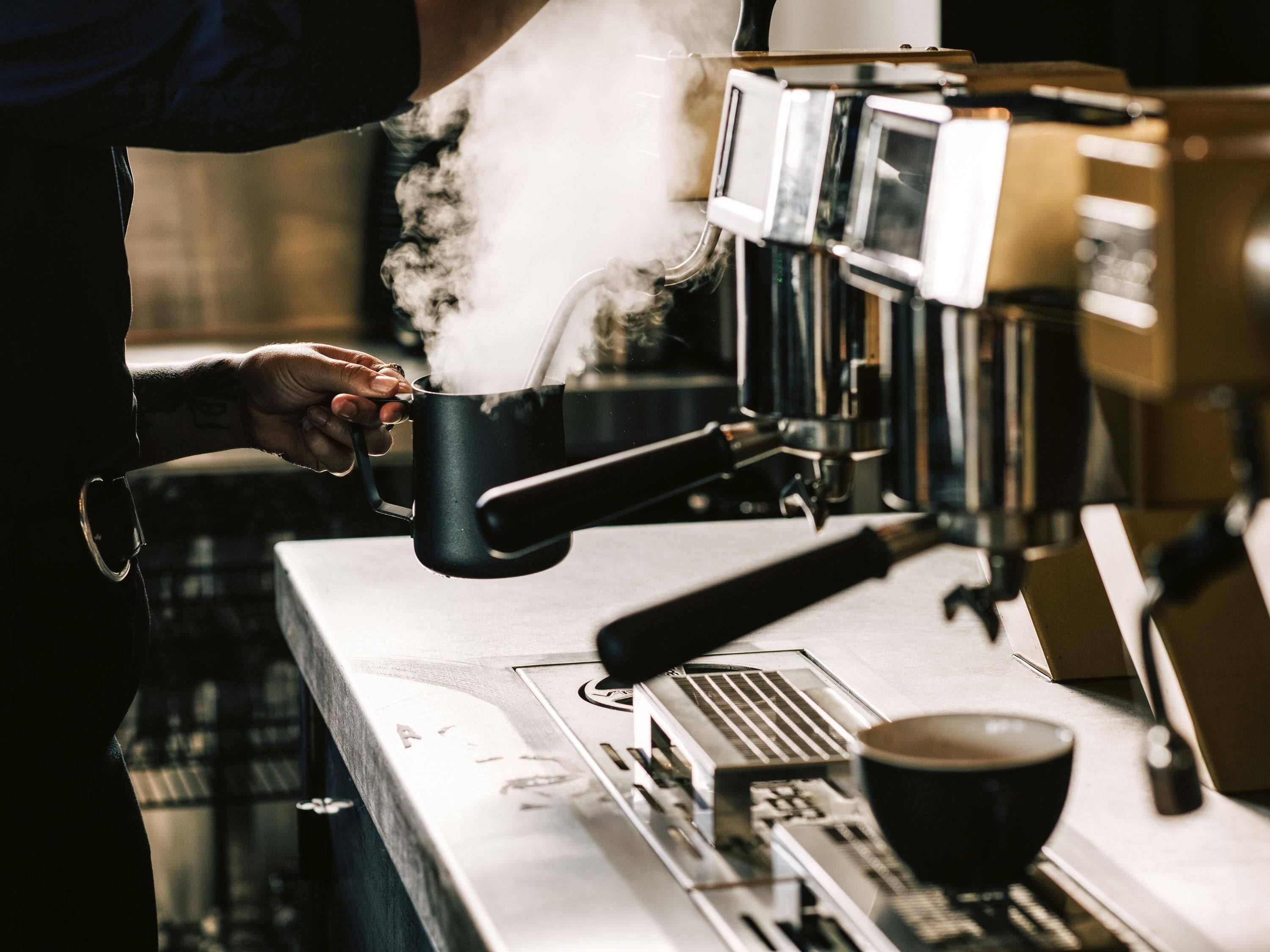 En person holder en kop nær en kaffemaskine, damp stiger op fra koppen, og en kaffekop står på disken.