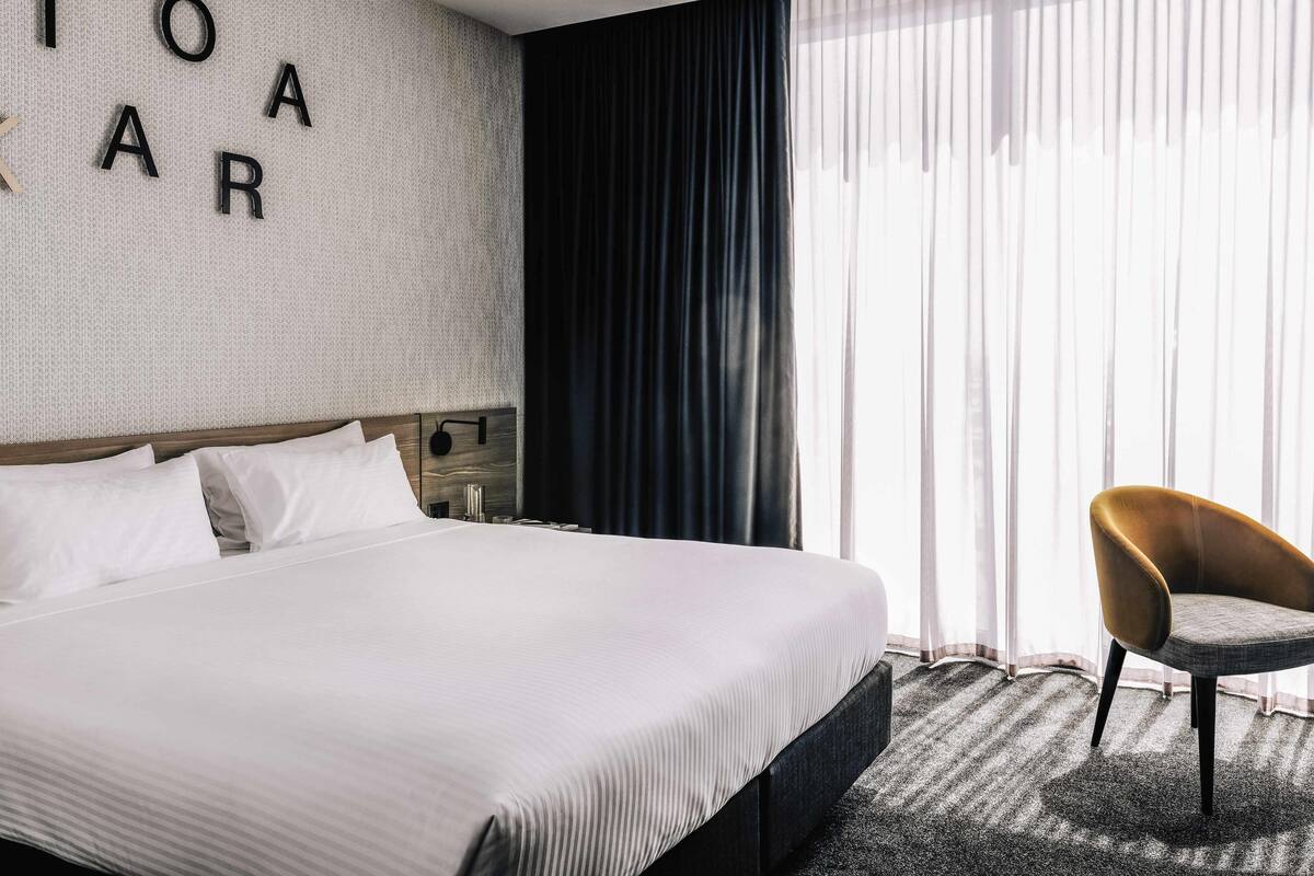 Ein Hotelzimmer mit einem großen Bett, einem Kopfteil mit Buchstaben, einem Nachttisch, einem Sessel und einem Fenster mit Vorhängen.