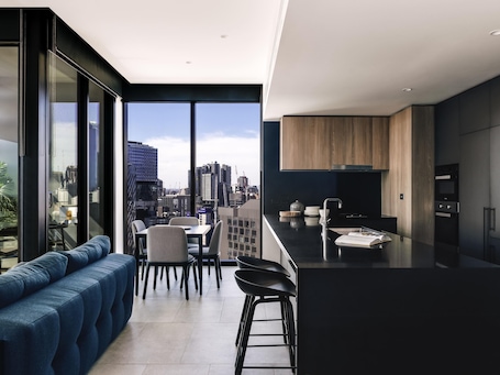 A modern kitchen with a city view, a dining area with a blue sofa, and a large window.. Novotel Melbourne South Wharf