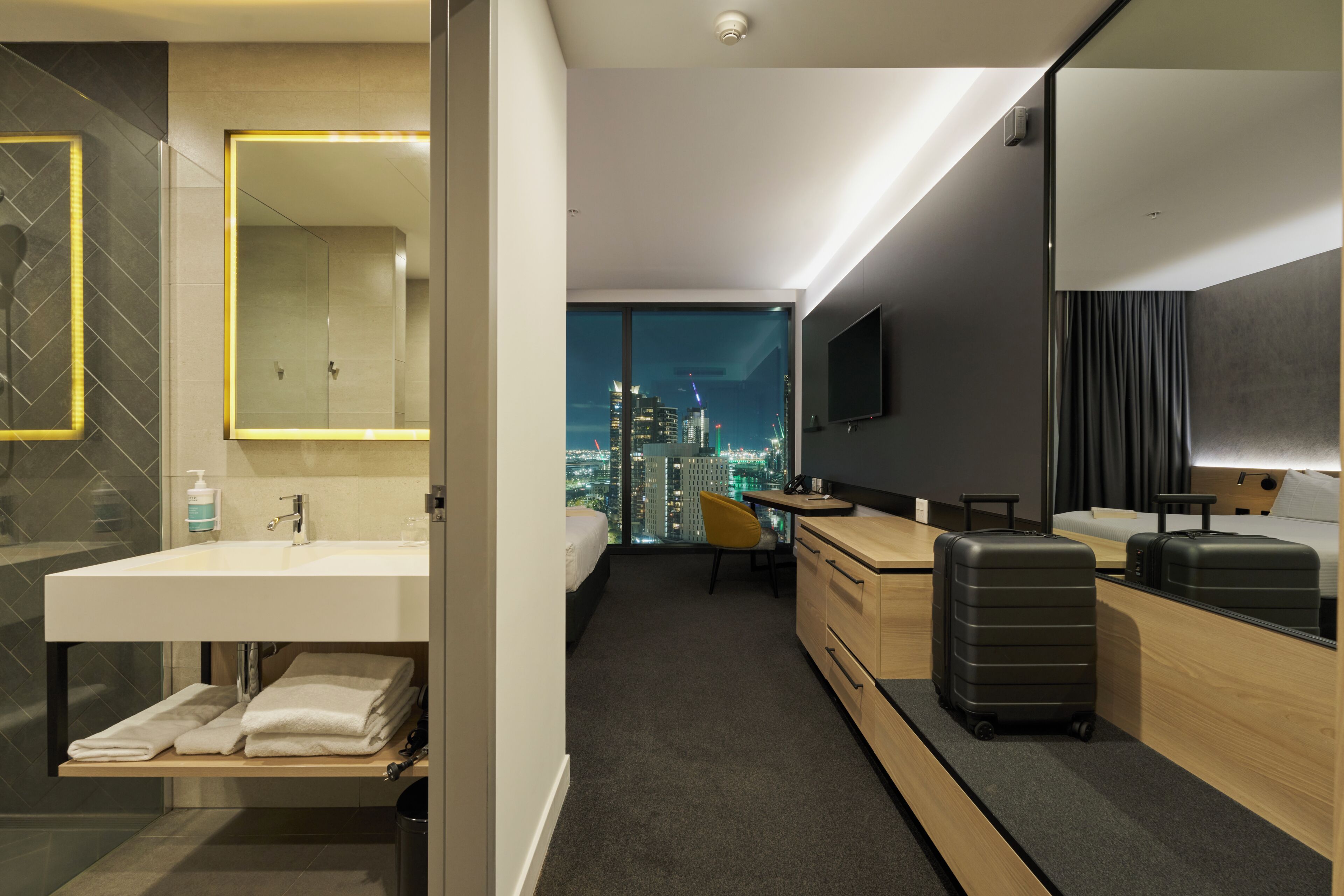A modern hotel room with a large window, a desk, a bed, a bathroom with a mirror, and a suitcase.