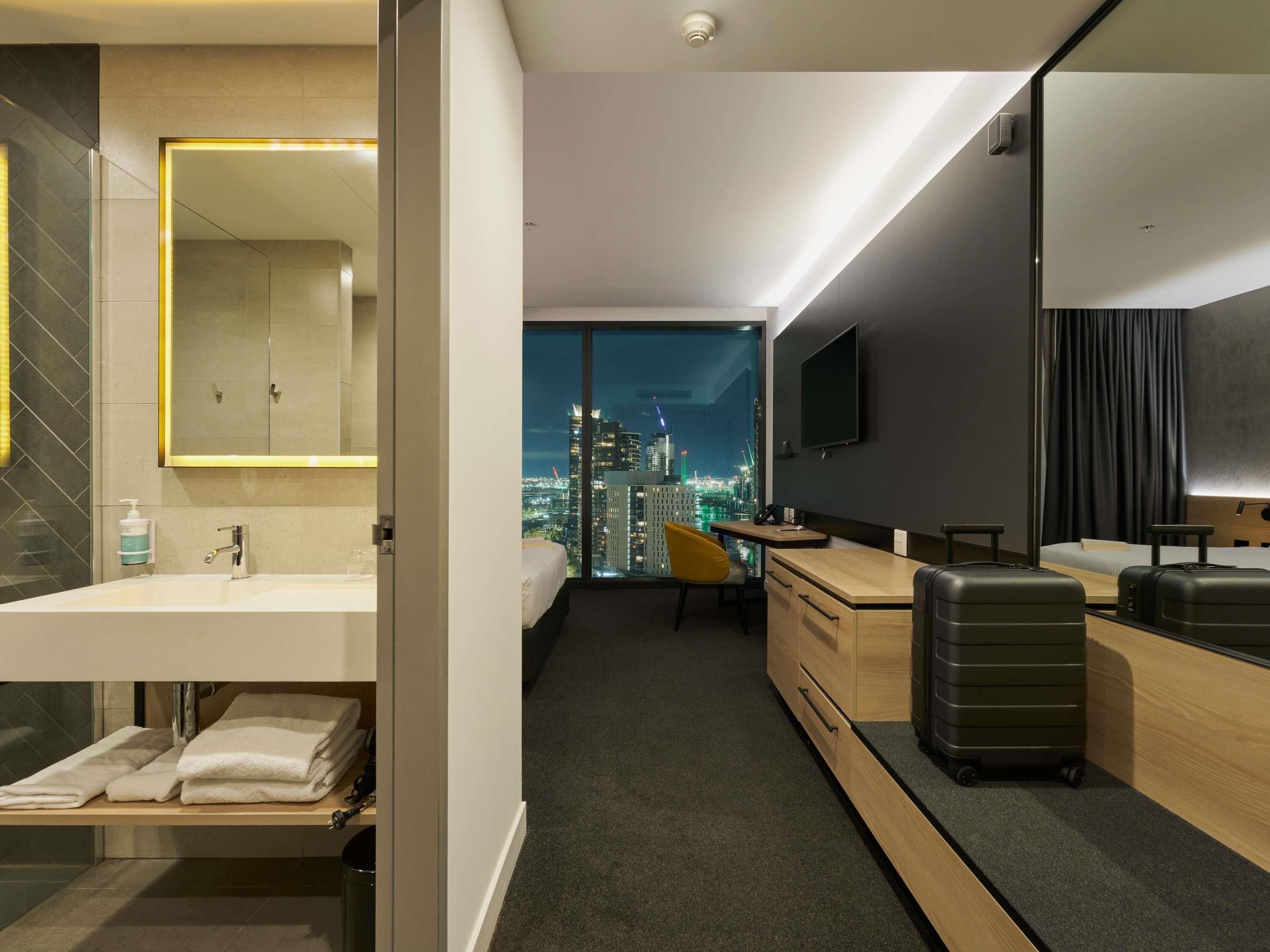 Une chambre d’hôtel moderne avec vue sur la ville, comprenant un bureau, une chaise, un lit, une télévision et une salle de bain avec un miroir et un lavabo.