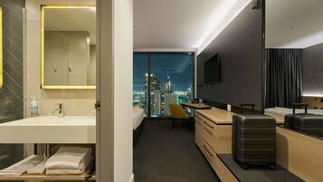 Une chambre d’hôtel moderne avec vue sur la ville, comprenant un bureau, une chaise, un lit, une télévision et une salle de bain avec un miroir et un lavabo.
