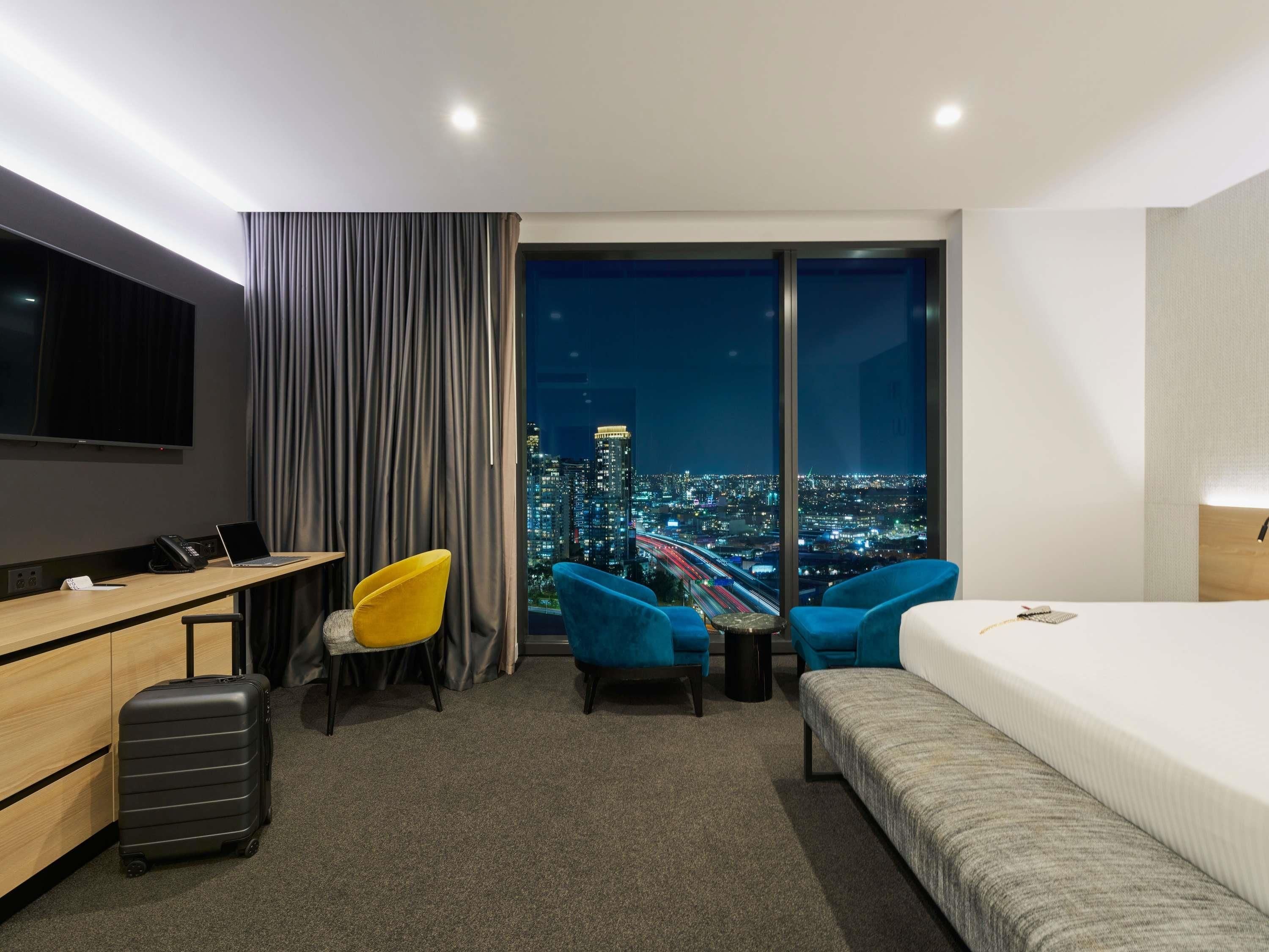 A hotel room with a bed, a desk, two chairs, a suitcase, a TV, and a city view.