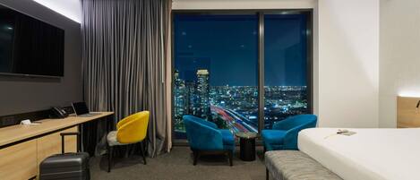 A hotel room with a bed, a desk, two chairs, a suitcase, a TV, and a city view.