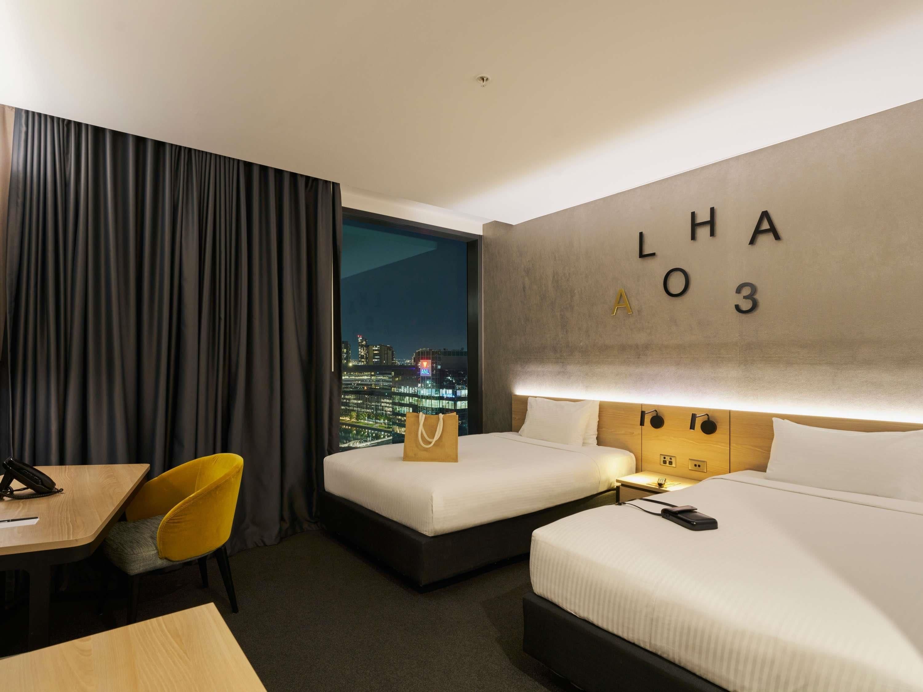 A hotel room with two beds, a desk, a chair, a view of the cityscape, and a wall with room number and letters.