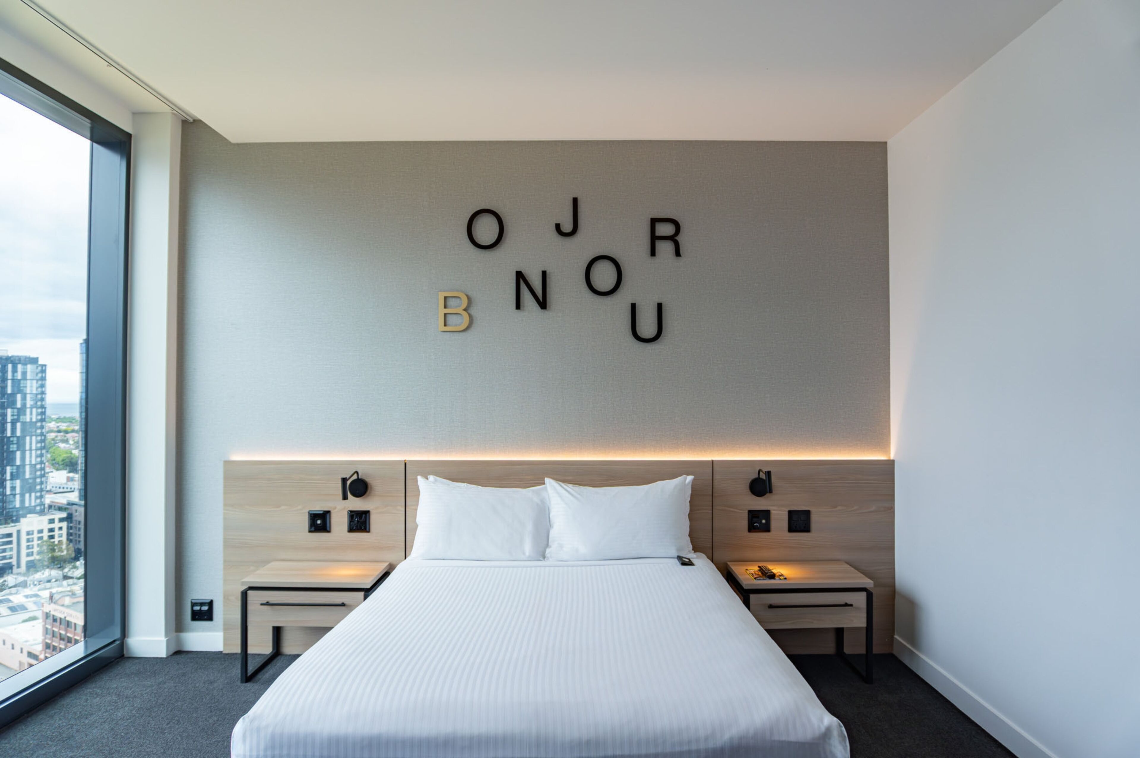 Quarto de hotel moderno com uma cama grande, duas mesas de cabeceira, uma luminária fixada na parede e vista para a cidade.