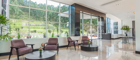 A spacious lobby with large windows, modern furniture, and a view of greenery outside.