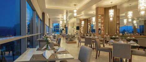 Un restaurante moderno con techos altos, amplias ventanas y candelabros. Las mesas están ordenadas con sillas.