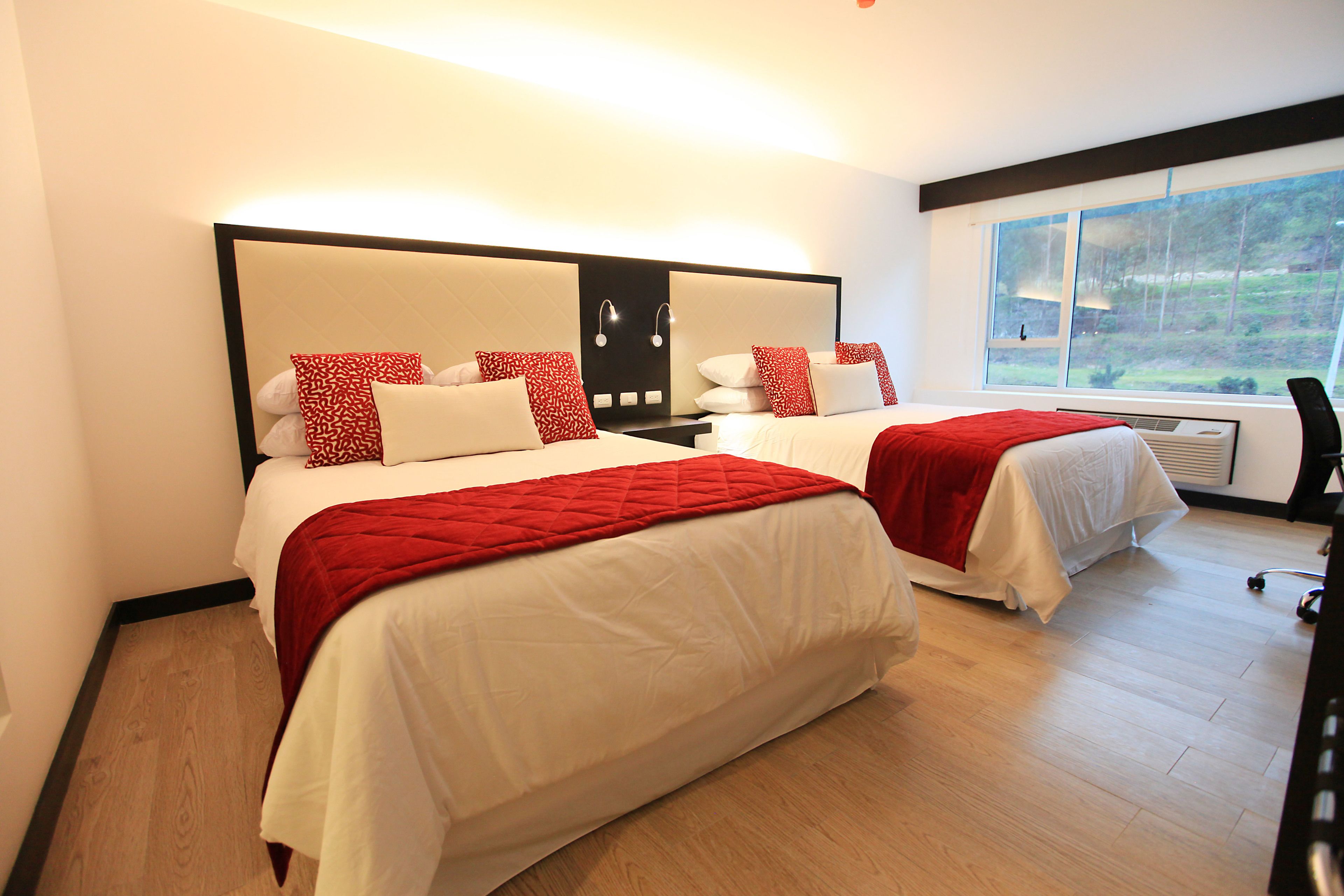 A hotel room with two beds, each with white bedding and red accents, a large window with a view of greenery, and a modern design.