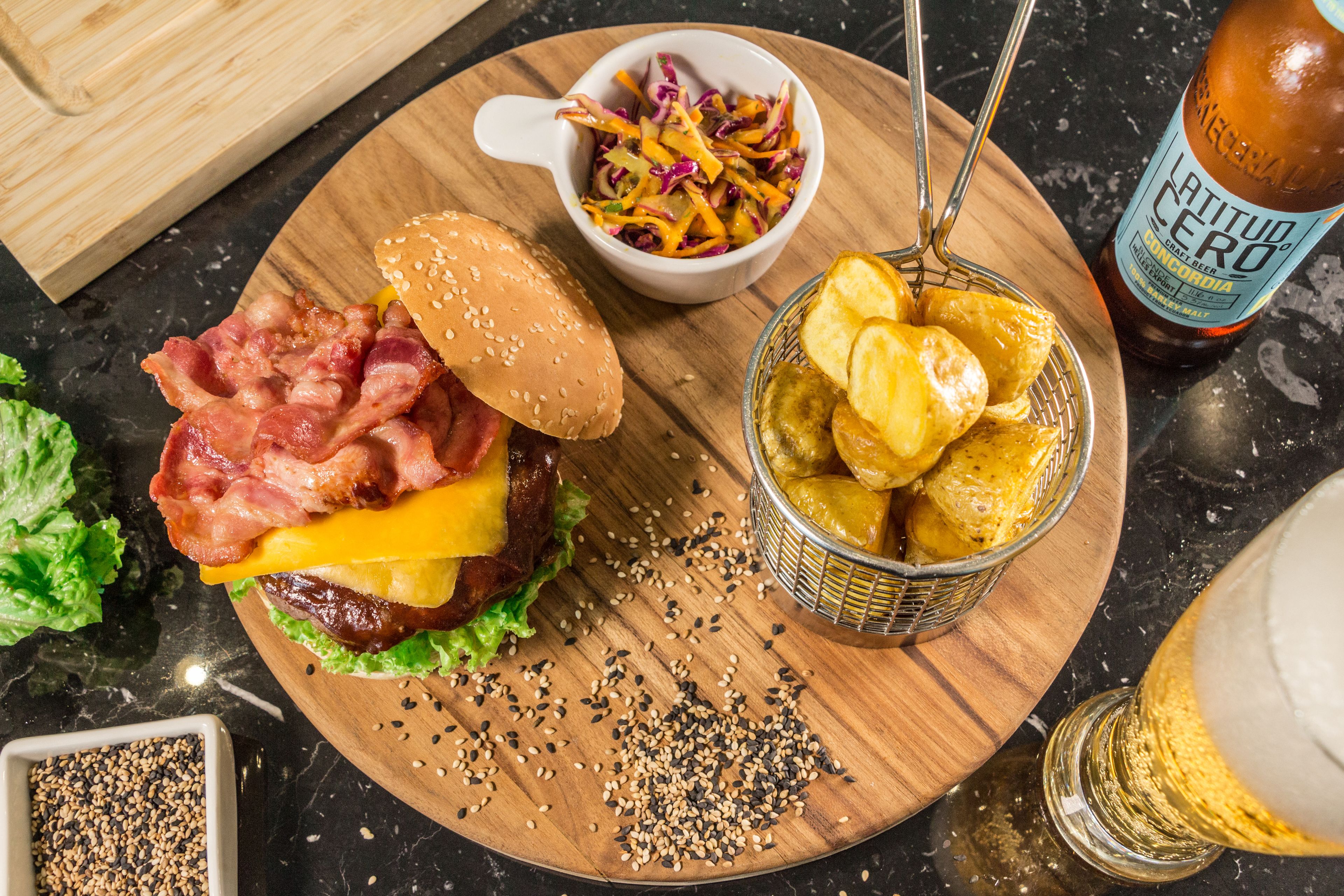 Una comida con hamburguesa, ensalada de col, papas fritas y una botella de cerveza sobre una tabla de madera.