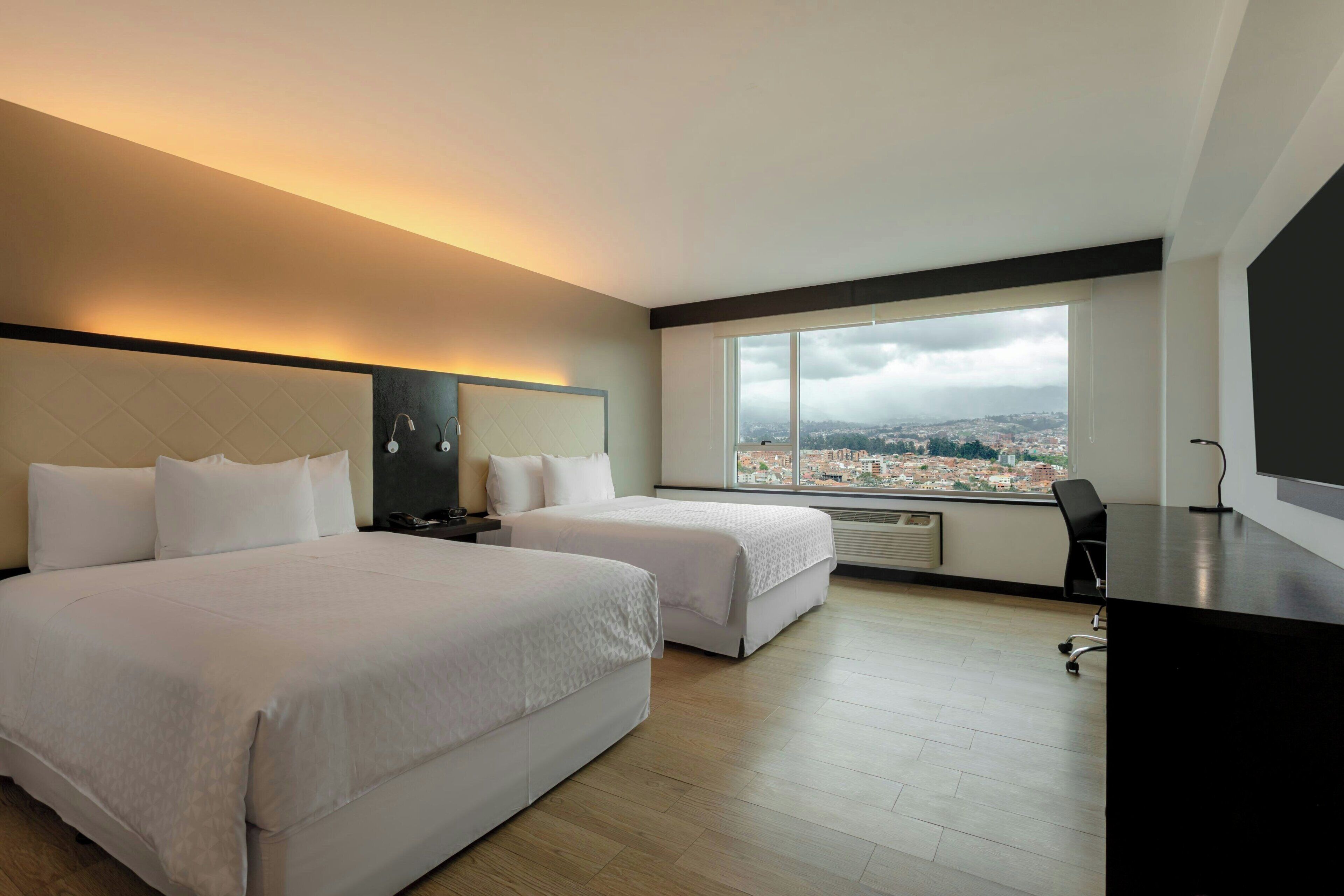 A hotel room with two beds, a desk, a chair, a large window with a city view, and a modern design.