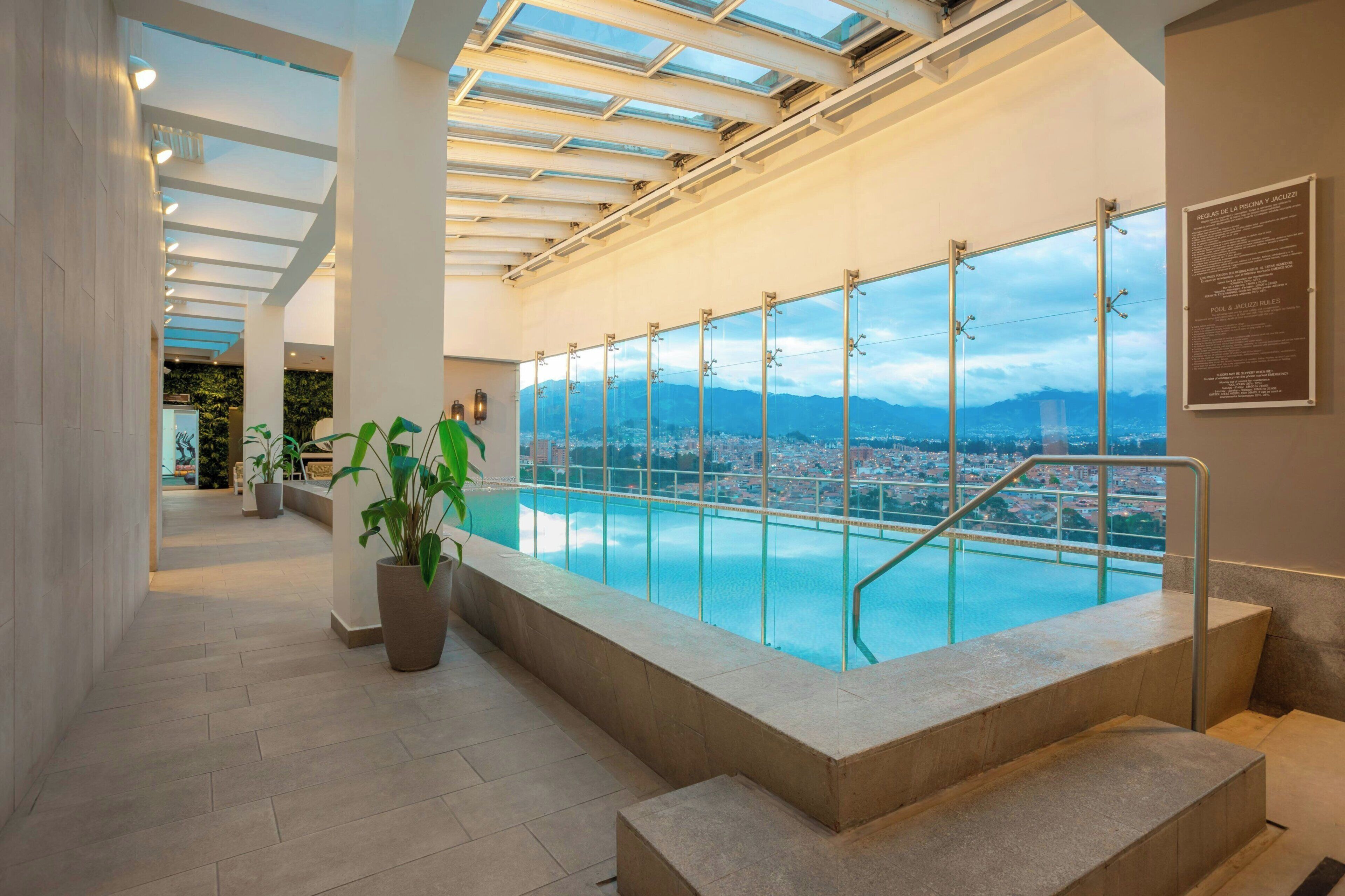 A modern indoor pool with a city view, a glass railing, and a staircase leading to the pool.