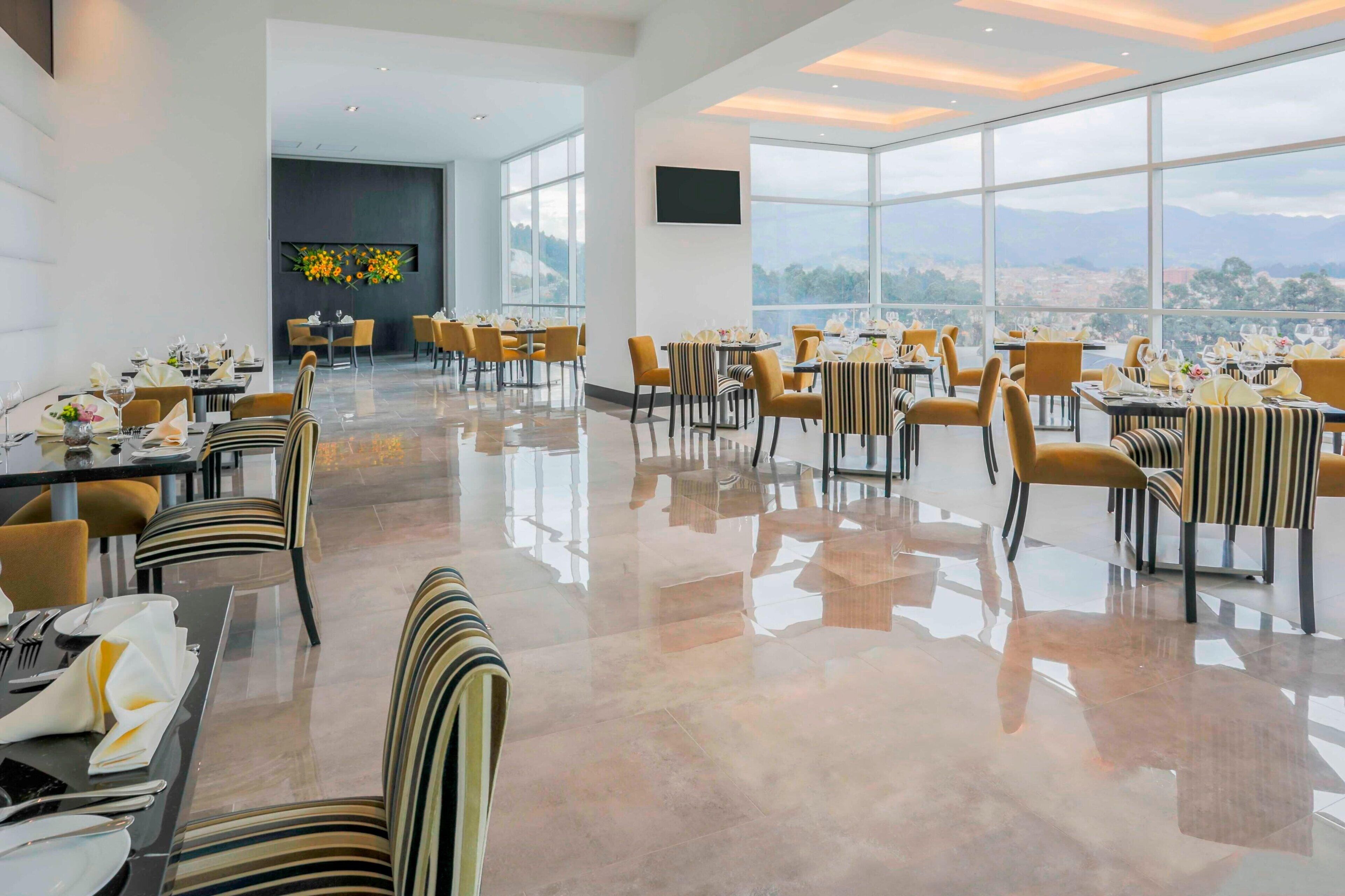 Un comedor amplio con mesas y sillas dispuestas para los huéspedes, ventanas grandes que ofrecen vistas al paisaje urbano, y un diseño de interiores moderno.