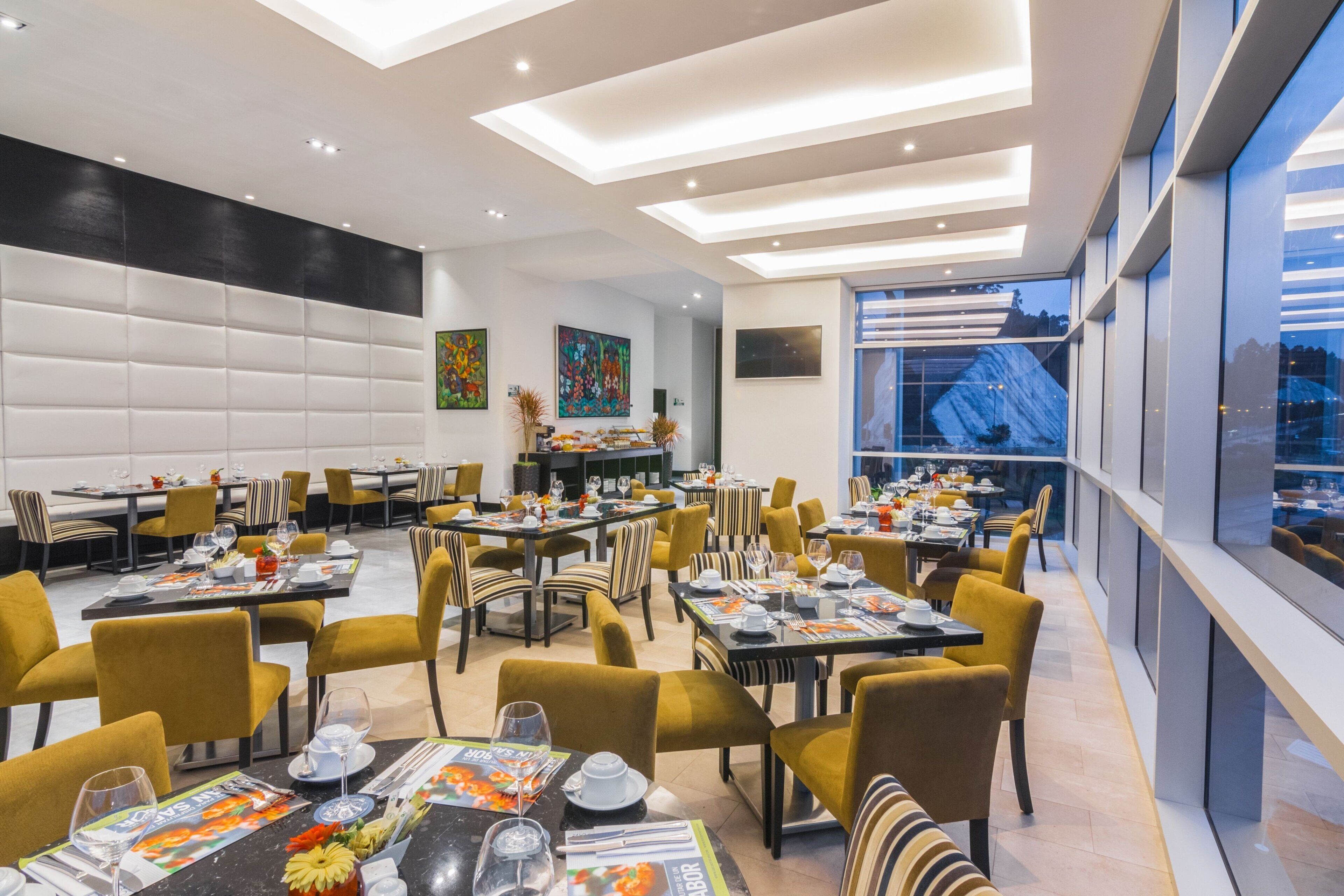 Een moderne eetruimte met meerdere tafels en stoelen, een hoog plafond met inbouwverlichting en grote ramen met uitzicht op de stad.