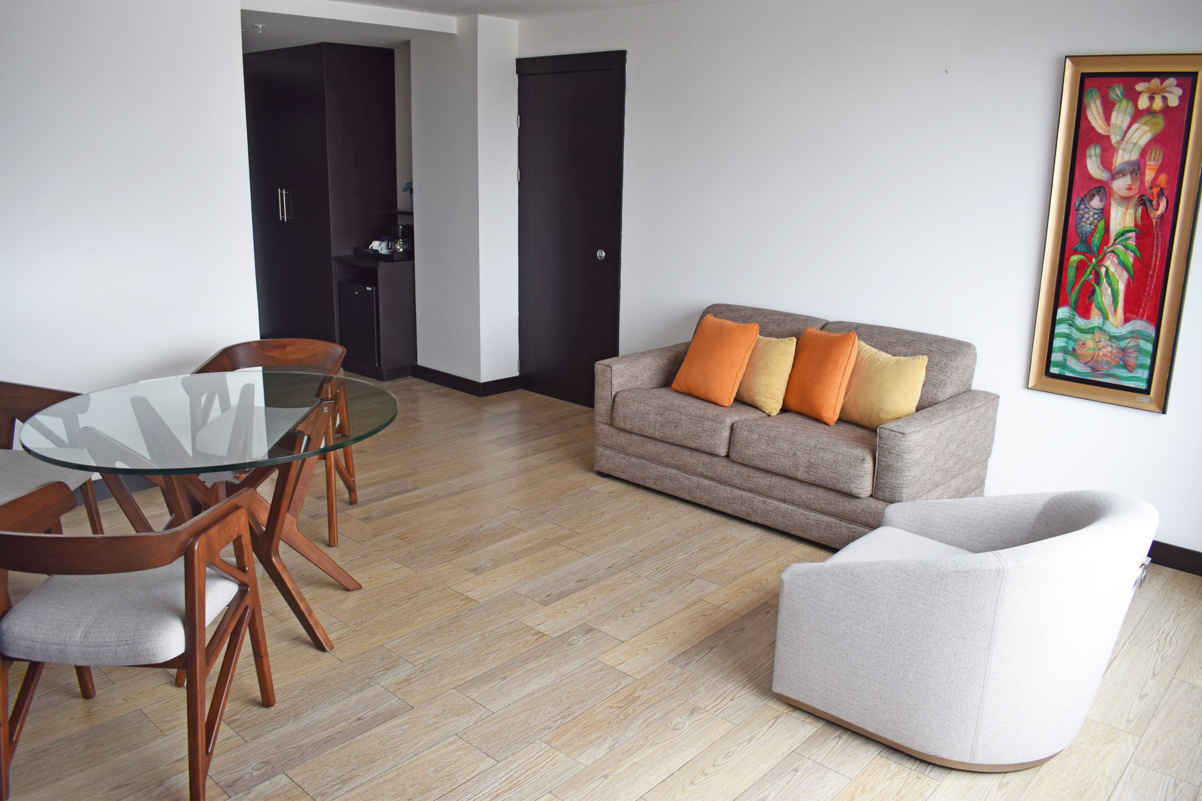 Ein modernes Wohnzimmer mit einem runden Glastisch, Holzstühlen, einem grauen Sofa mit orangefarbenen Kissen und einem weißen Sessel.