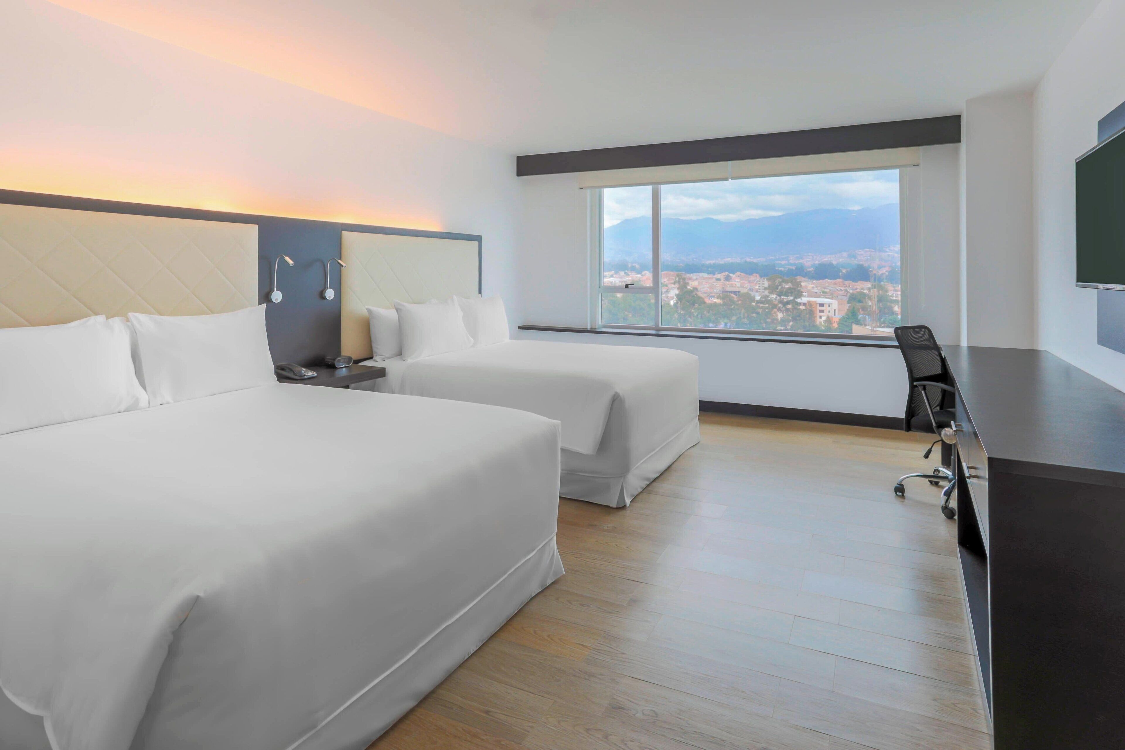 Habitación de hotel con dos camas, un escritorio y un ventanal amplio con vistas a la ciudad.
