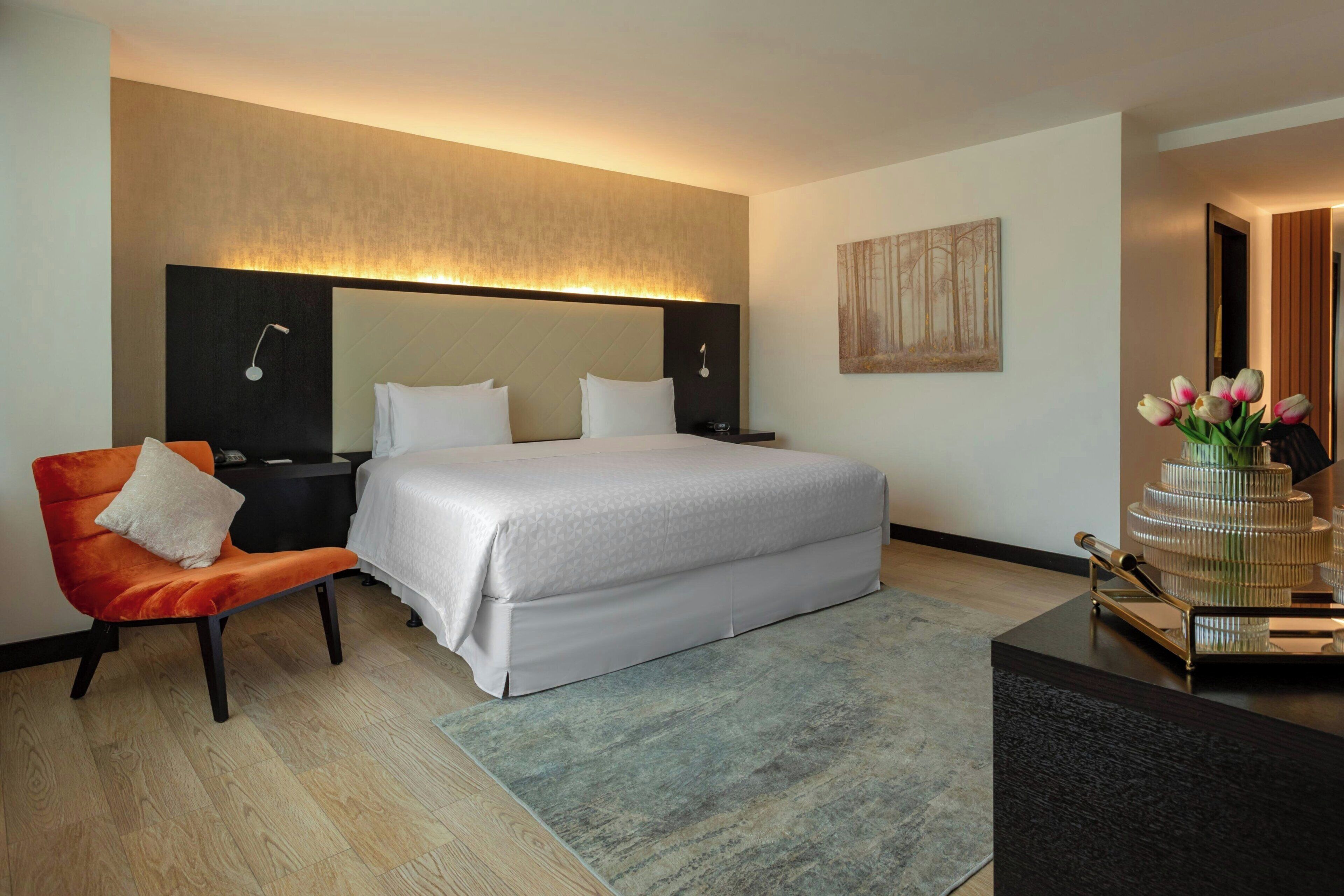Una habitación de hotel con una cama, una silla, una mesita de noche, un espejo, una obra de arte en la pared y un florero con flores.