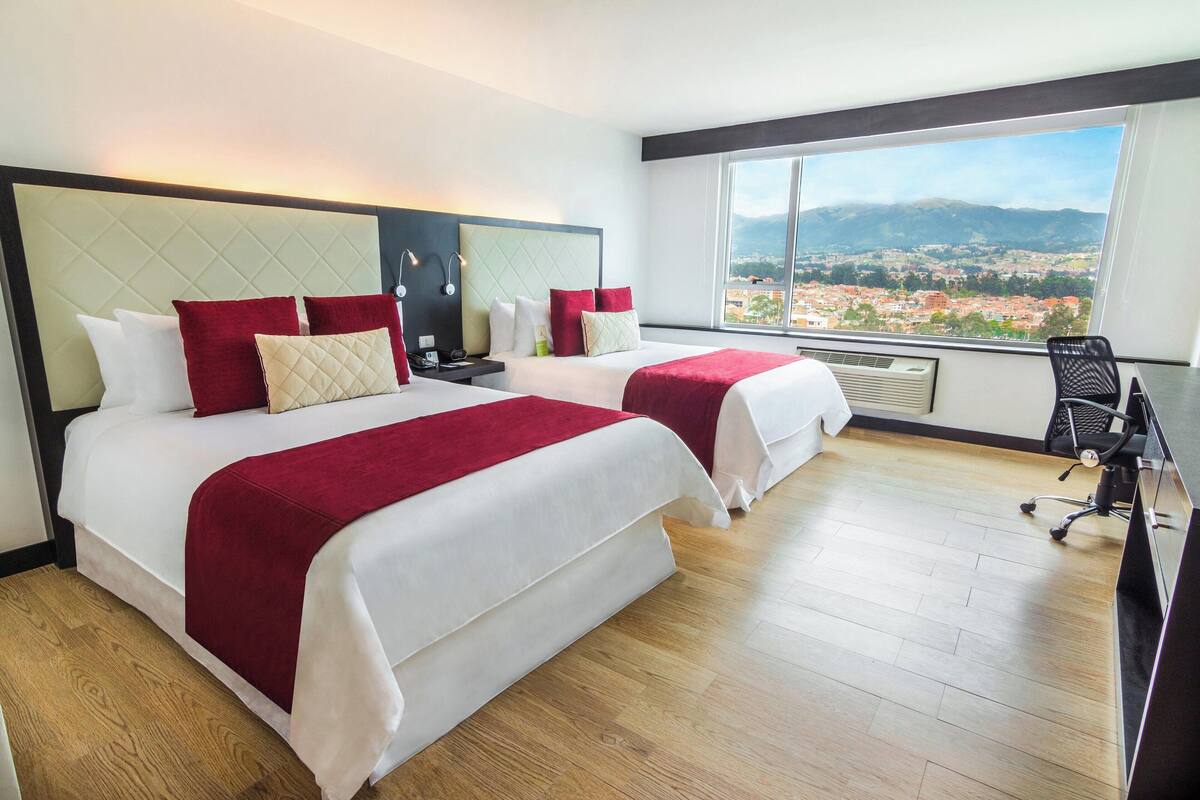 Habitación de hotel con dos camas, un escritorio y un amplio ventanal con vistas a la ciudad.