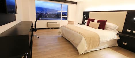 A modern hotel room with a large bed, a desk, and a view of a cityscape through a large window.