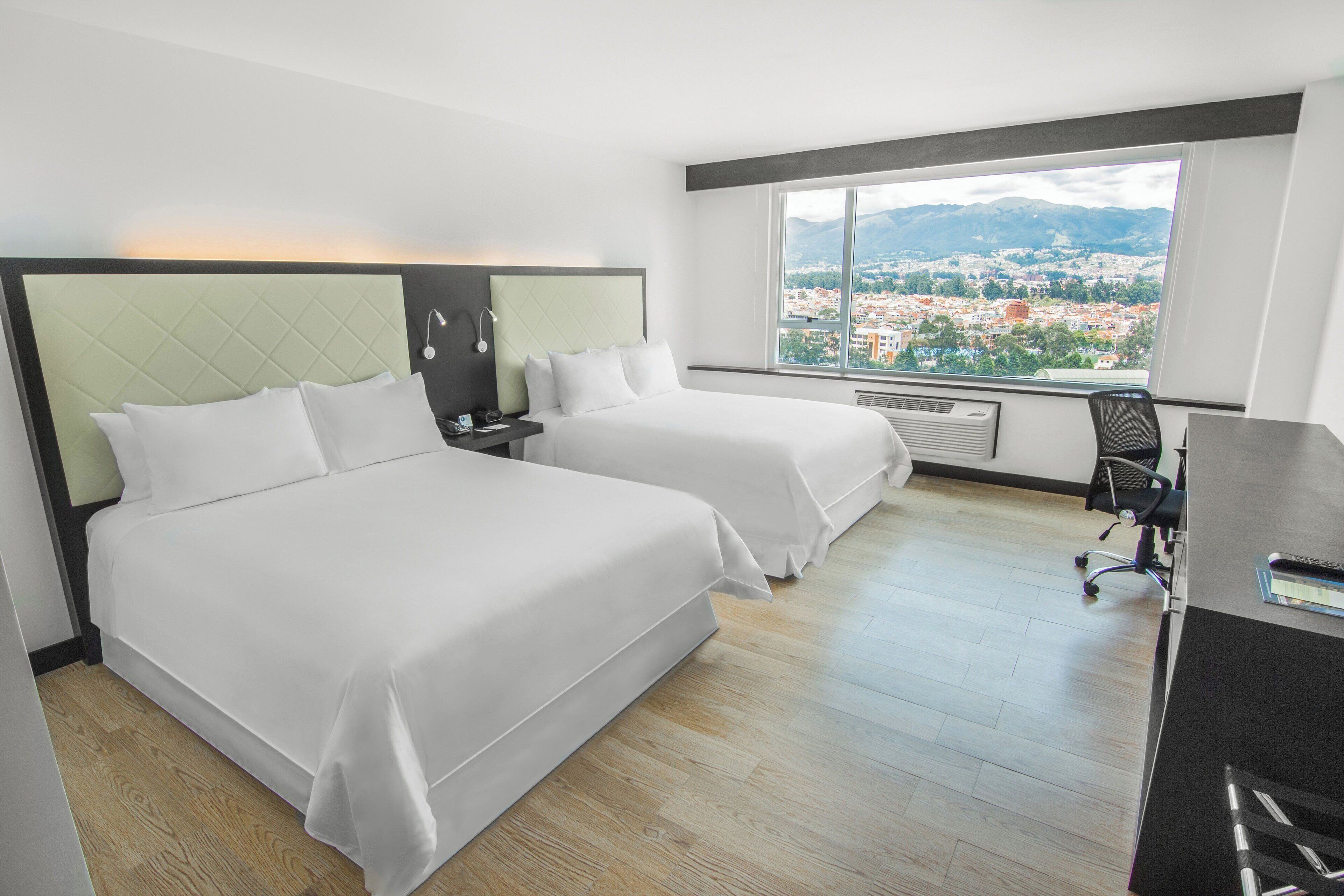 A hotel room with two beds, a desk, a chair, and a large window offering a city view.