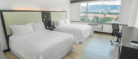 A hotel room with two beds, a desk, a chair, and a large window offering a city view.