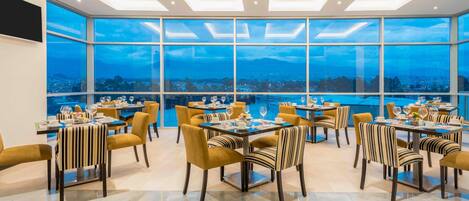 A dining area with multiple tables and chairs, large windows offering a city view, and a modern ceiling design.