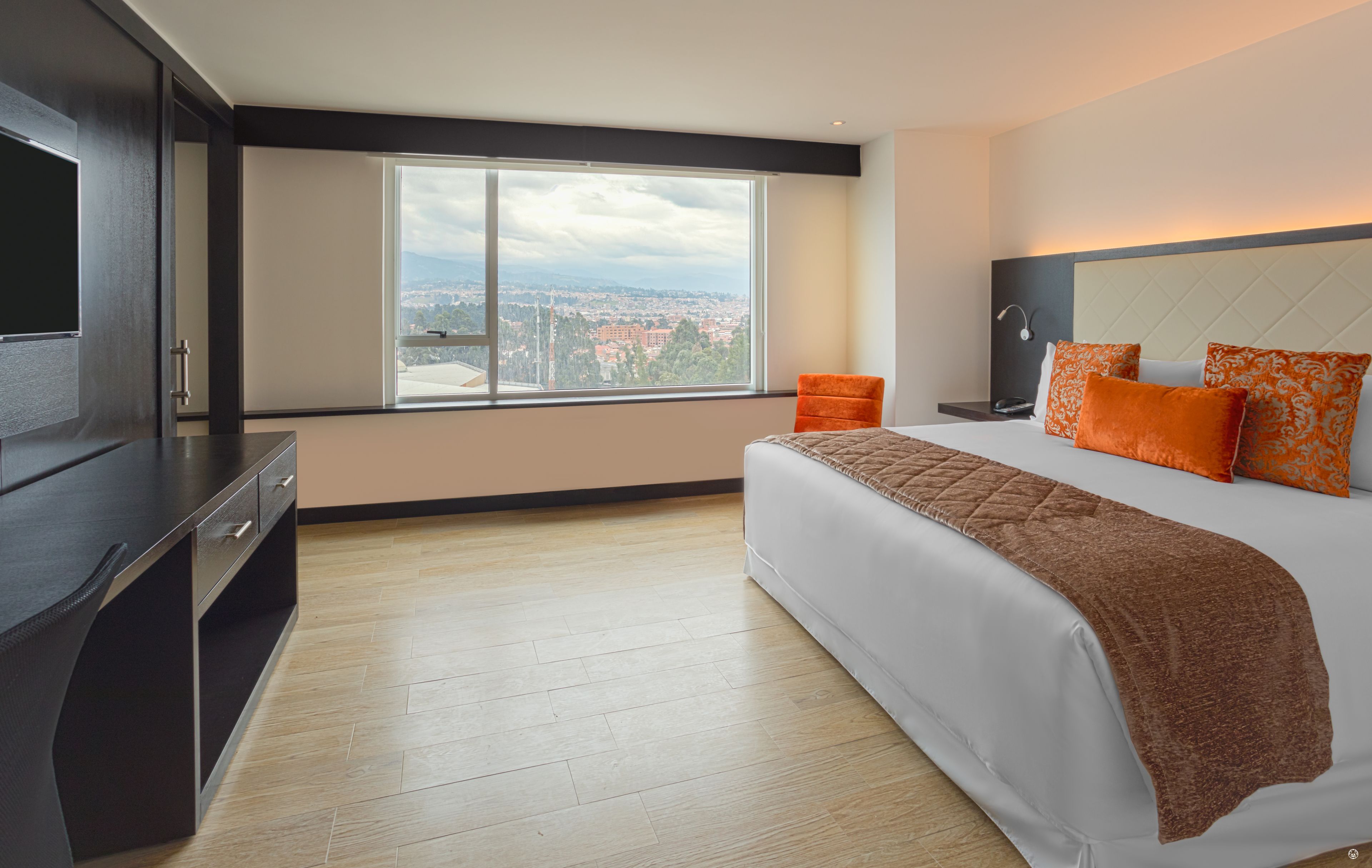 A modern hotel room with a large bed, a TV, a desk, and a view of a cityscape.