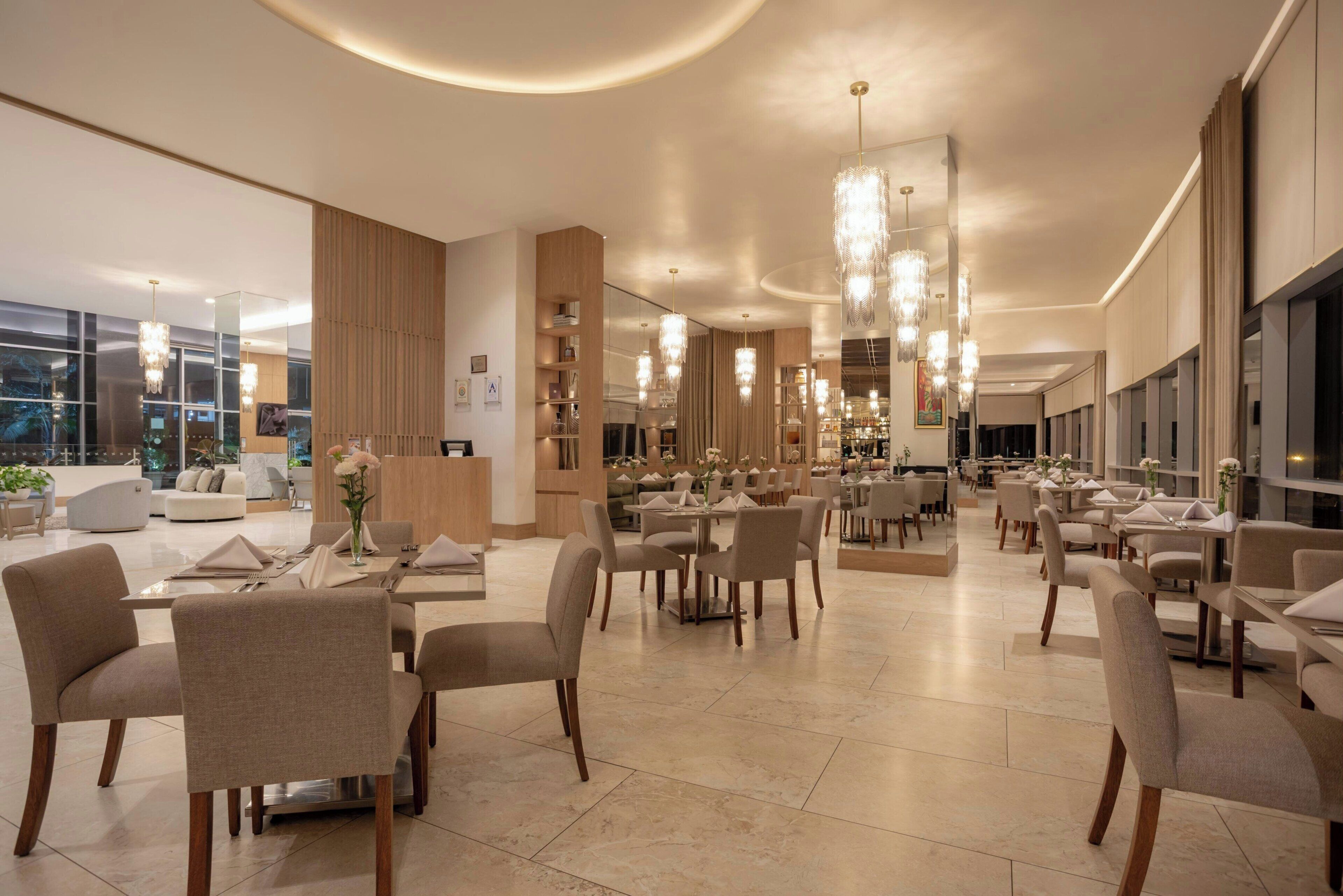 Ampla área de refeições de hotel, com várias mesas e cadeiras, janelas grandes e lustres modernos.