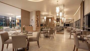 Ampla área de refeições de hotel, com várias mesas e cadeiras, janelas grandes e lustres modernos.