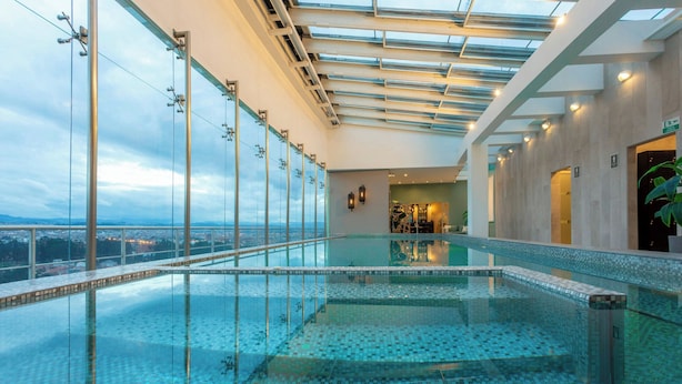 Une piscine à débordement entourée de parois en verre, offrant une vue sur la ville, dotée d’une verrière et présentant un aménagement intérieur moderne.