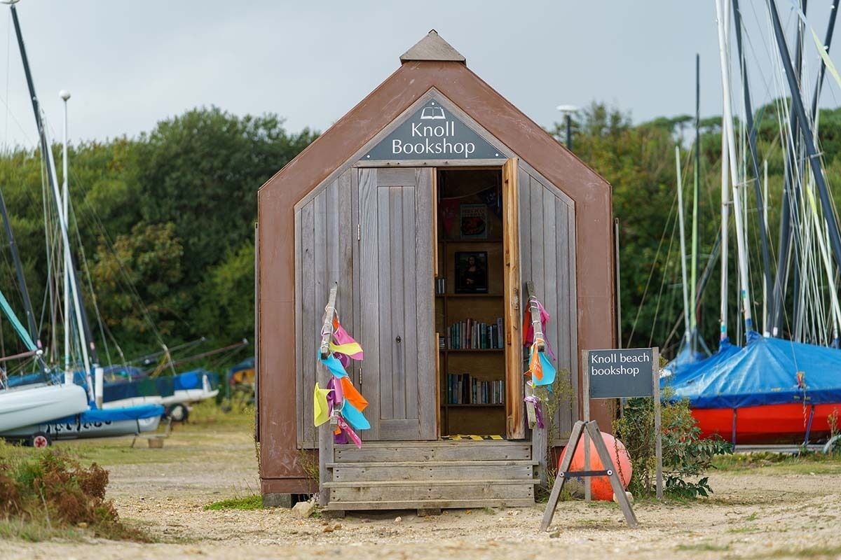 A wooden Knoll Bookshop with a triangular roof, located near a beach with sailboats.