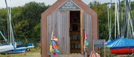A wooden Knoll Bookshop with a triangular roof, located near a beach with sailboats.