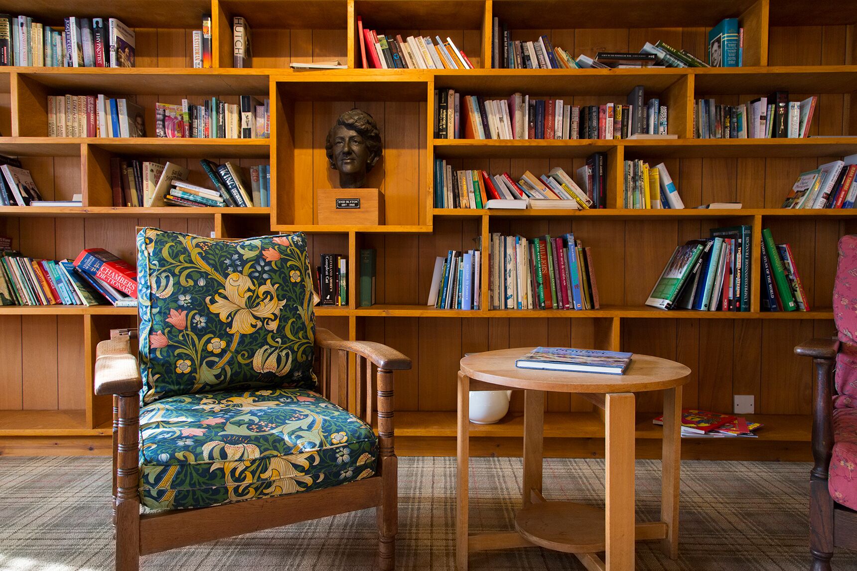 Una biblioteca con estanterías de madera llenas de libros, un sillón con cojín floral y una pequeña mesa de madera.