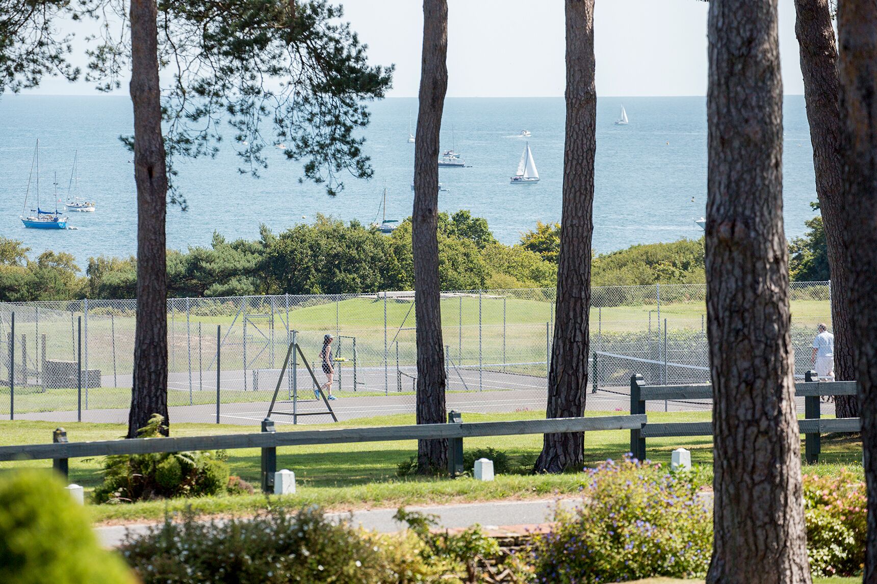 A view of a tennis court with a fence, trees, and sailboats on the water in the background.