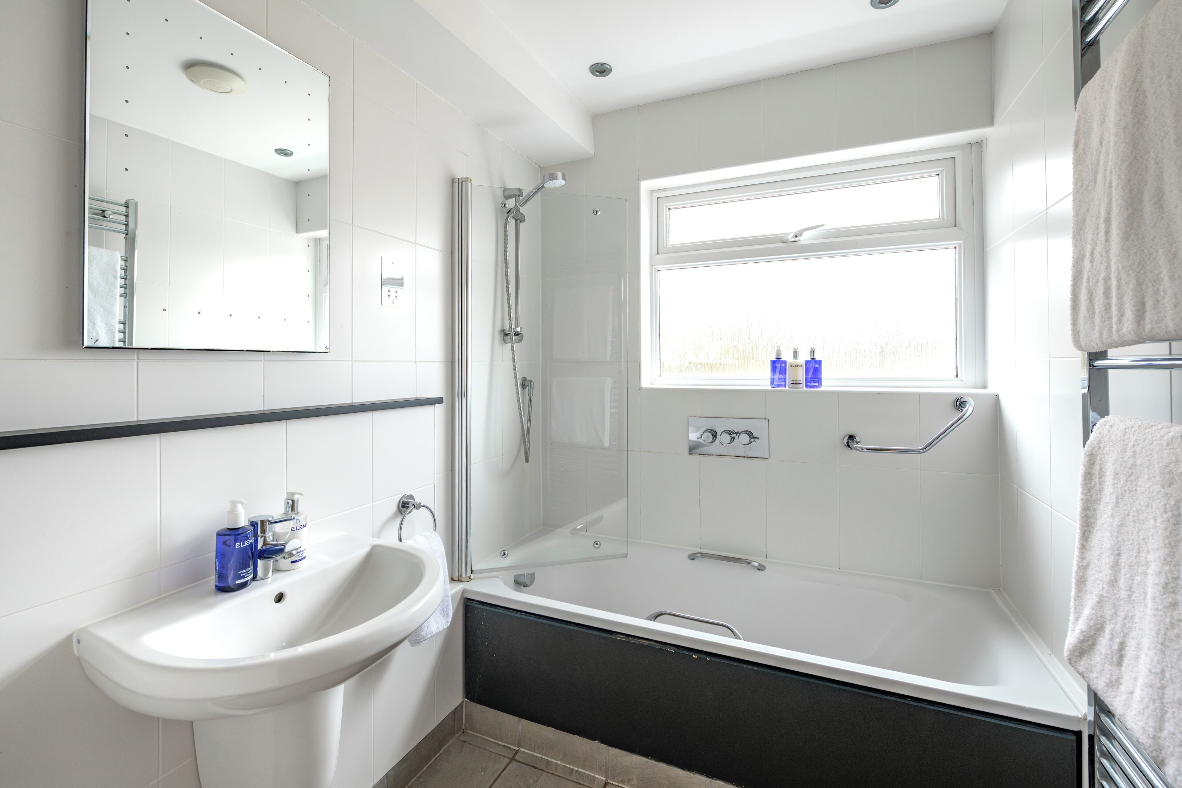 Een moderne badkamer met een wit wastafelmeubel, een badkuip, een douche en een raam.