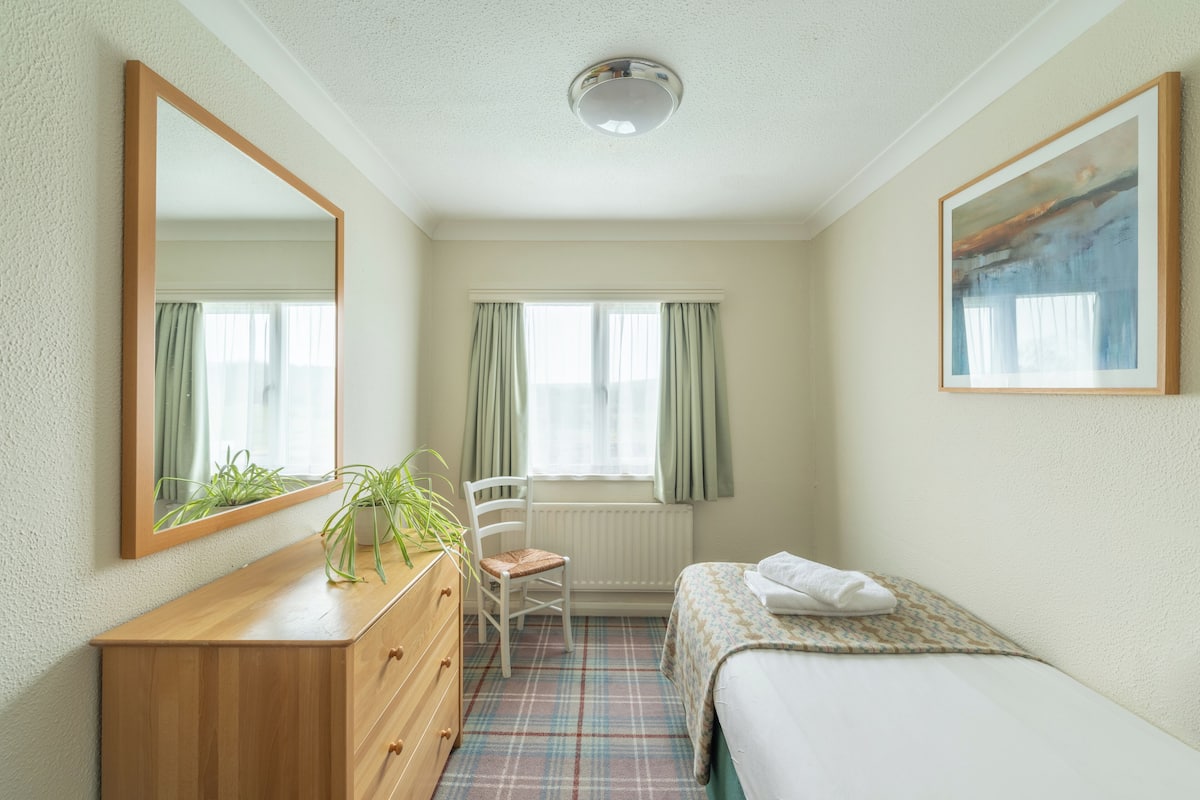 A bedroom with a bed, a dresser, a chair, a radiator, a window with curtains, and a painting on the wall.