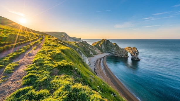 A coastal landscape with a grassy path leading to a beach, a rocky outcrop, and the ocean. - The Knoll House (Swanage)