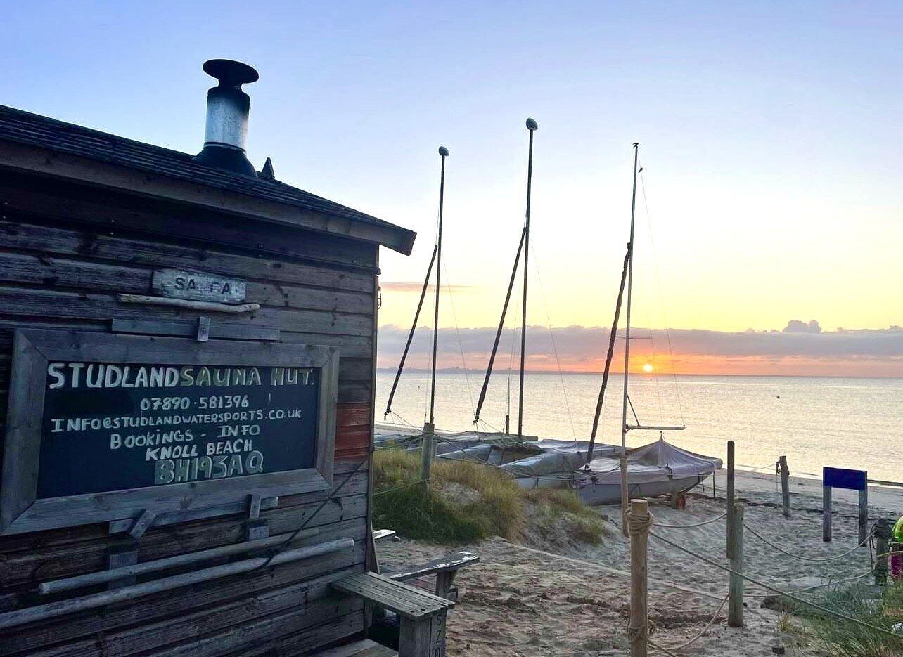 A wooden building with a signboard for Studlands Sauna Hut, including contact information and booking details.